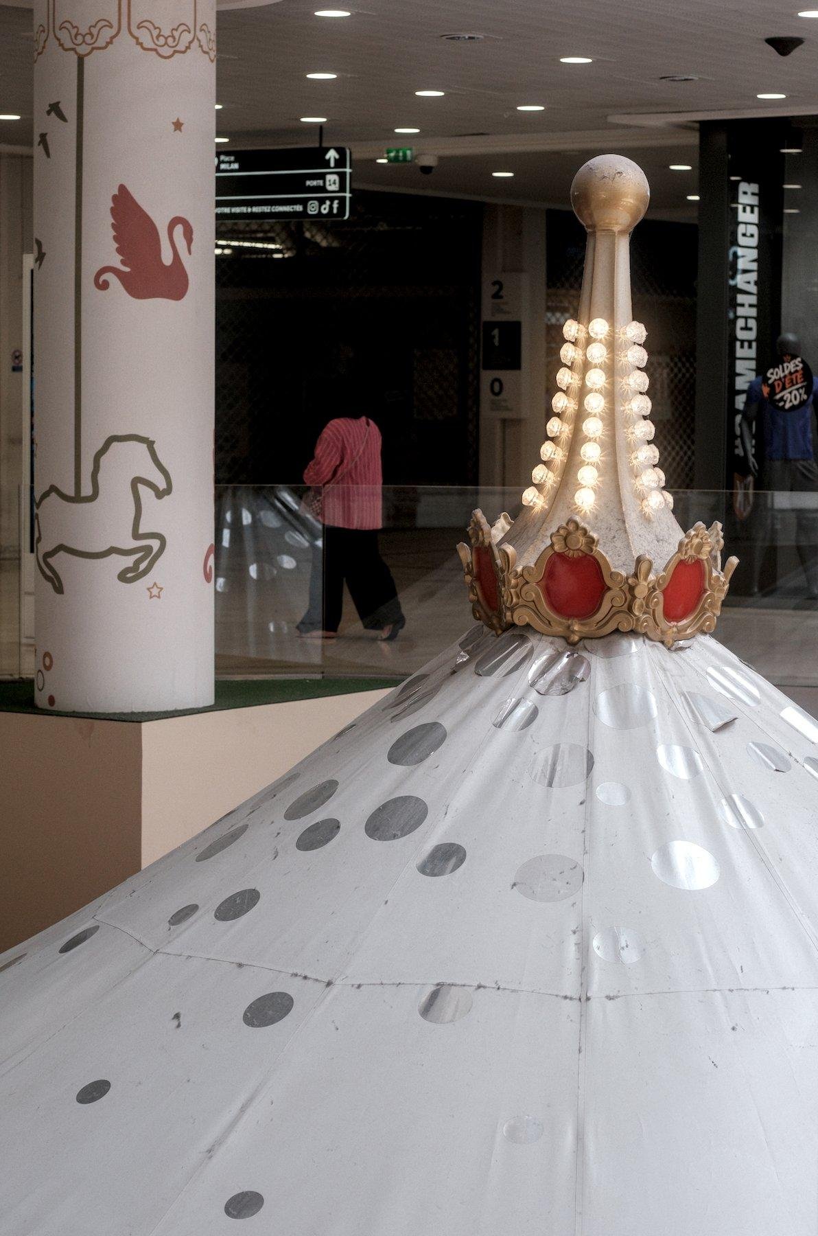 Photo verticale. Dans une galerie marchande, au premier plan, décalé à droite, le sommet d’un manège, blanc à pois argenté, avec le sommet décoré d’une pointe doré et rouge avec des petites lampes. À gauche, un poteau de soutiens, décoré avec des silhouettes de chevaux de manèges. Entre le poteau et la pointe, une dame en haut rouge et pantalon noir marche vers la gauche
