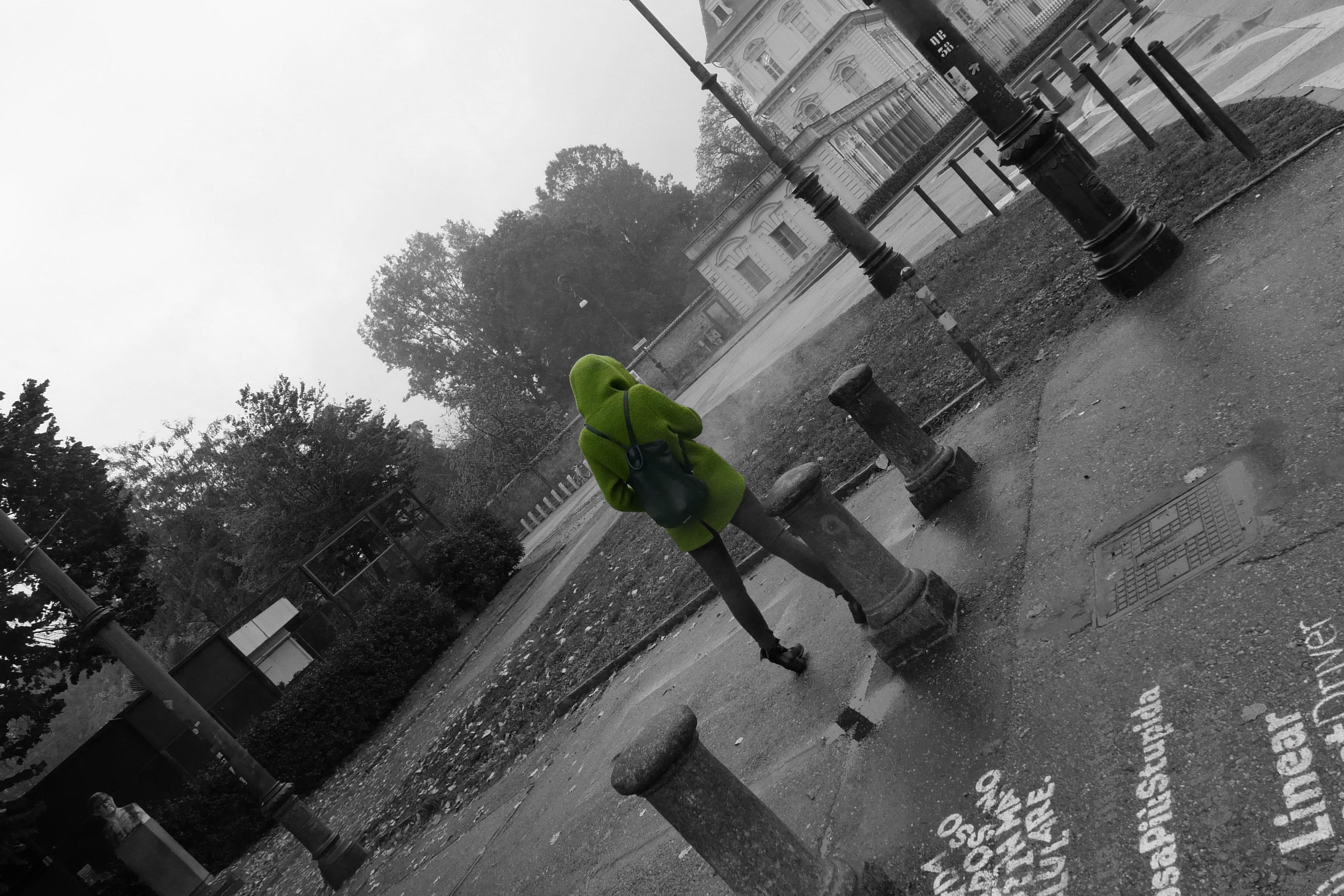 The image shows a person in a bright green outfit, walking on a street with several bollards. The person is the only element in color, while the rest of the image is in black and white. In the background, there are trees and a building. The ground has some text and patterns visible.
