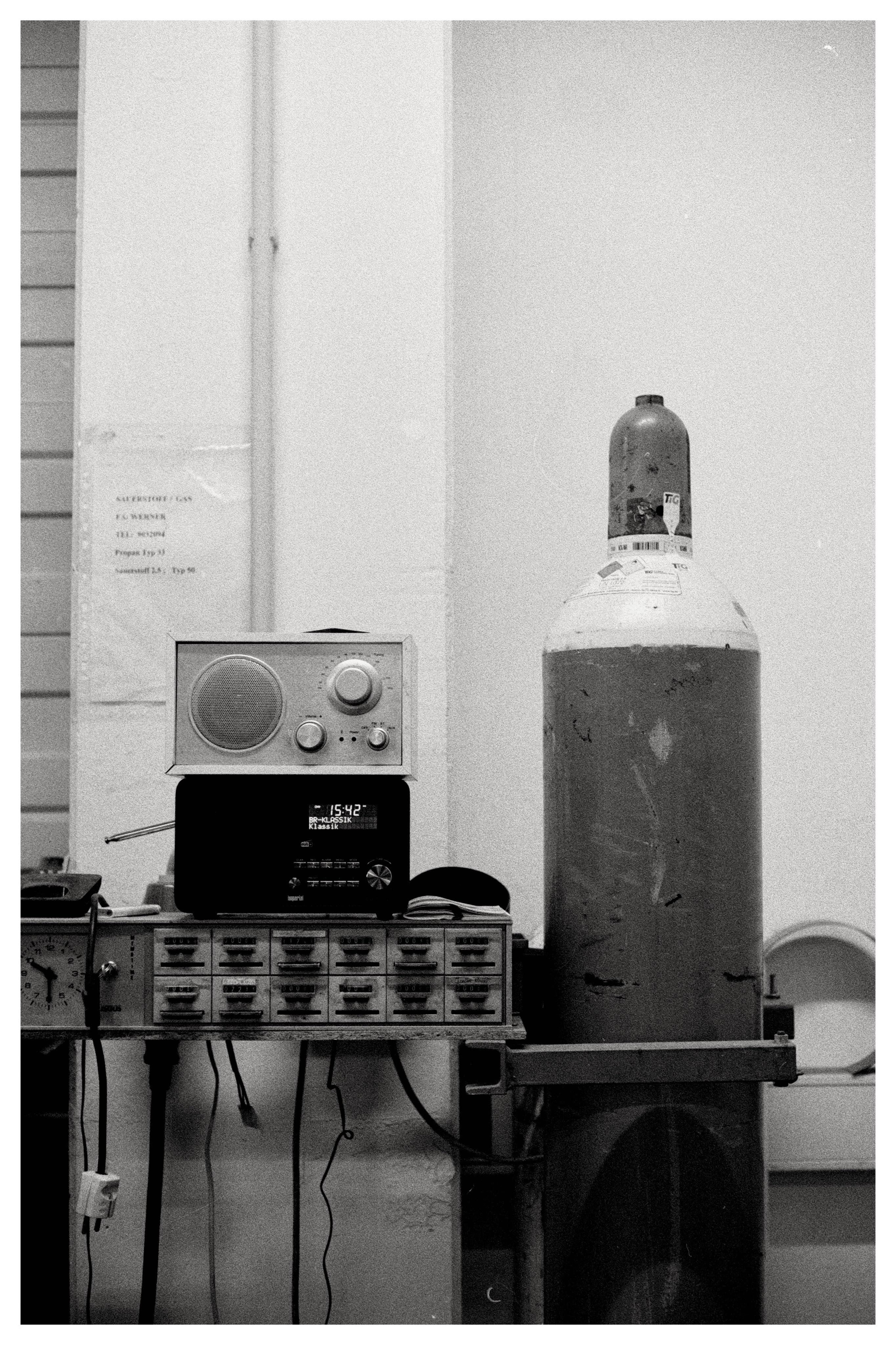 Black-and-white photo of a workbench with a small drawer cabinet, cords hanging below, and two radios stacked on top; a large gas cylinder stands to the right against a plain wall with a posted paper notice and vertical pipes in the background.