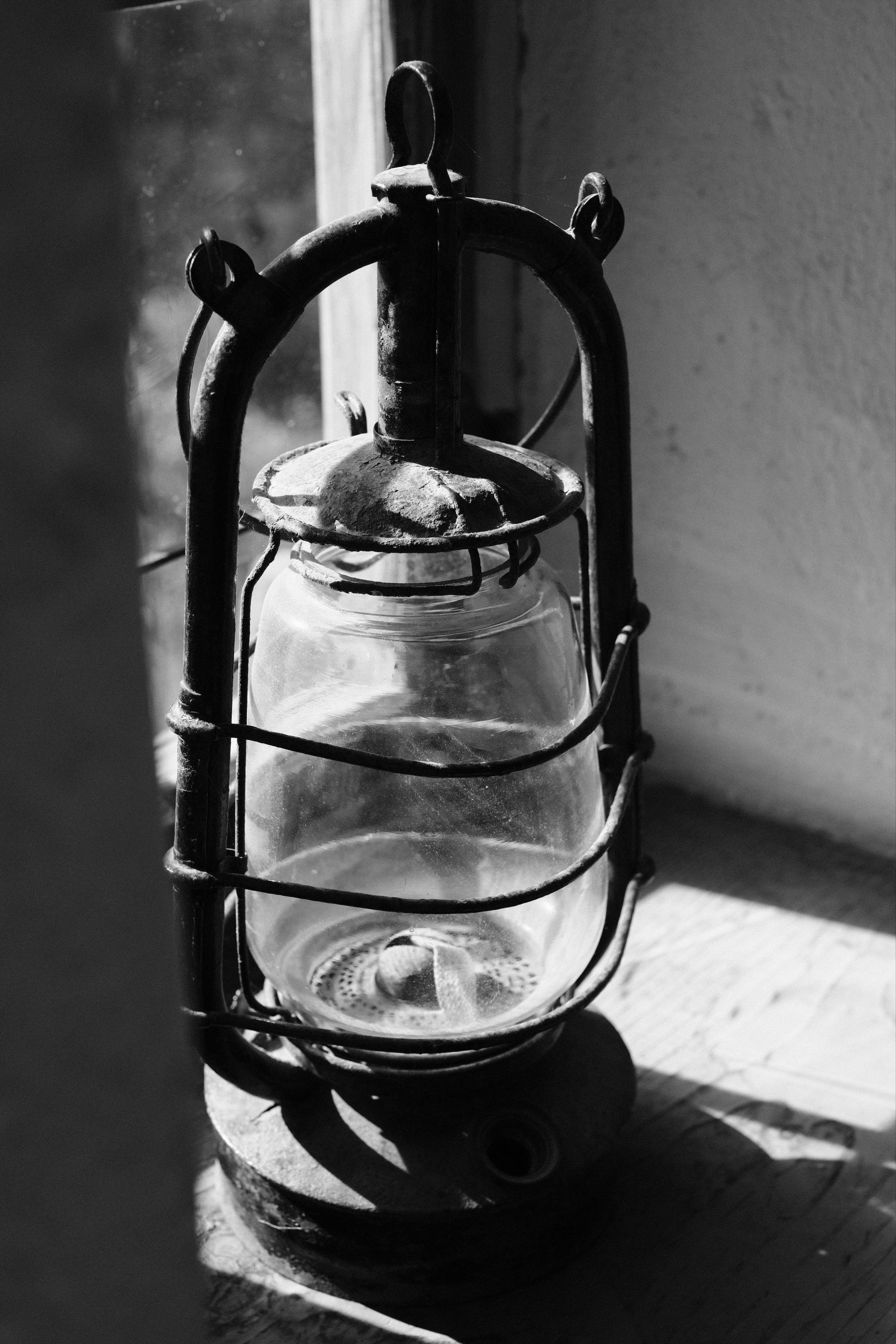 Black-and-white close-up of a vintage metal kerosene lantern with a glass globe, sitting on a wooden window sill. Harsh sunlight through the window casts strong shadows and contrasts.