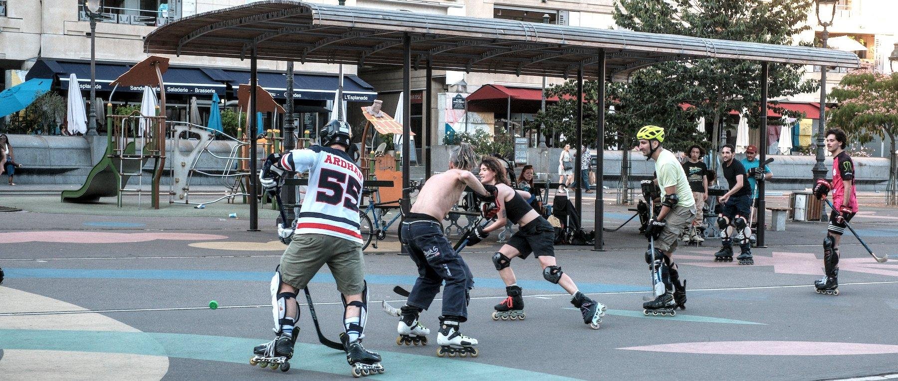 Photo horizontale. De jeunes gens en roller sur une place piétonne en train de jouer à du hockey sur bitume. Certains ont des casques, protège tibia, genouillère, coudière… Une jeune femme tente une percée et un homme se fait surprendre