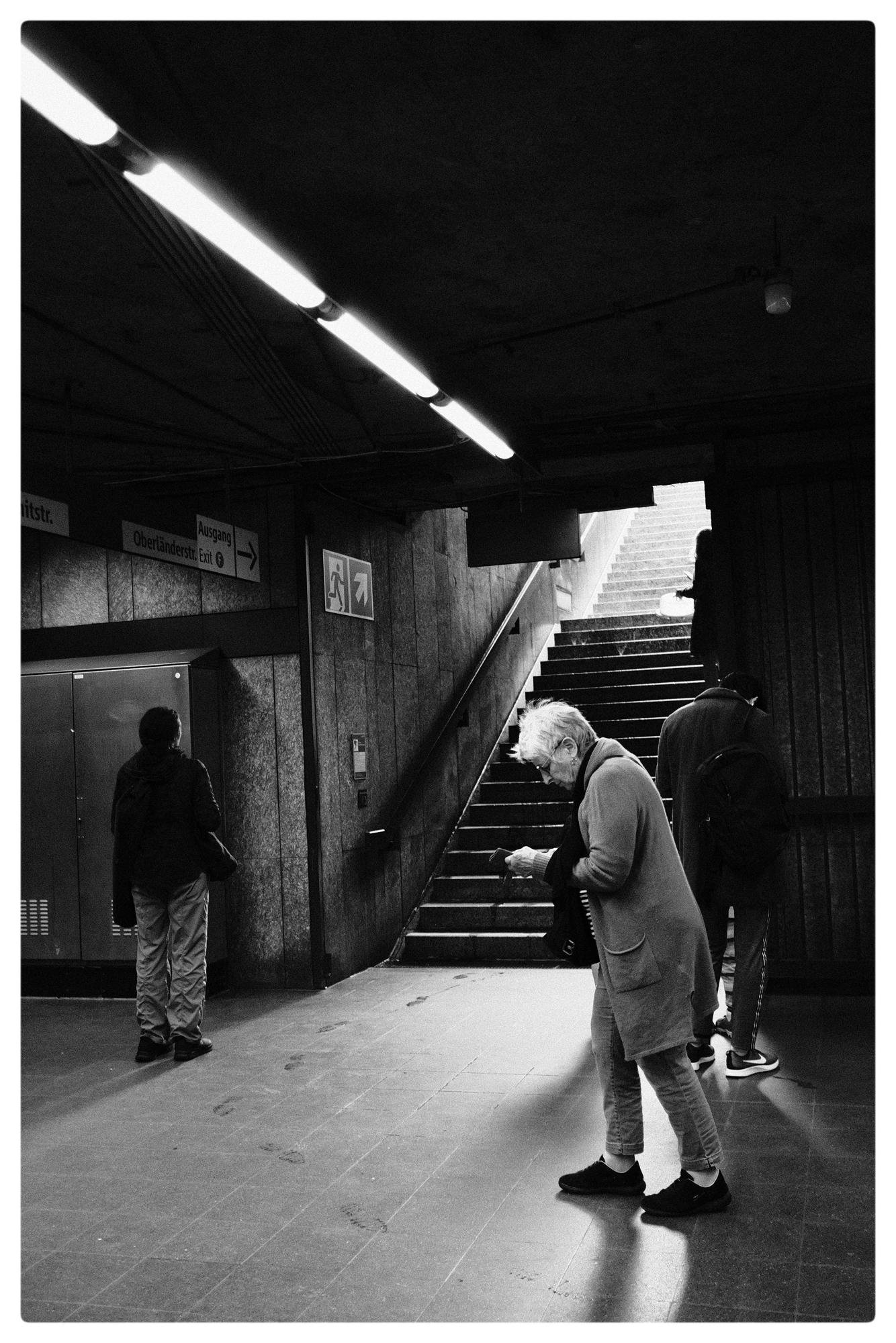Black and white photograph of a subway station interior with three people. One person is standing near a wall on the left, another is walking up a flight of stairs in the center, and an elderly person is checking something in their hand on the right. The ceiling has fluorescent lights, and signs above indicate directions with arrows labeled “Oberlandstr.” and “Exit”. The stairs lead up to a bright outdoor area. Footprints are visible on the floor.