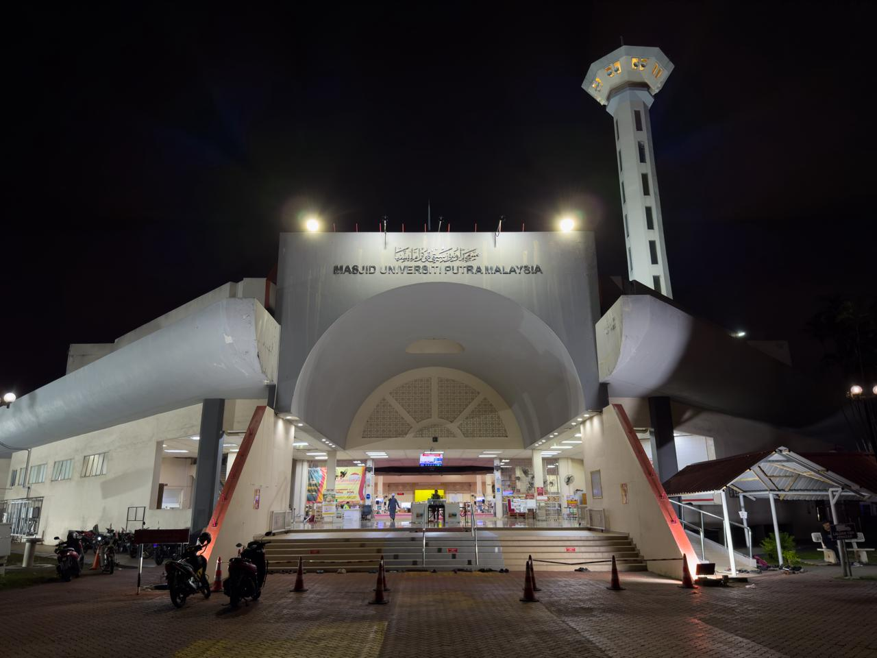 Night view of the entrance to the Universiti Putra Malaysia mosque. The facade is illuminated with large arches and a flat roof. A tall minaret stands on the right. The pathway leading to the entrance is lined with orange traffic cones and a few parked motorcycles. The sky is dark, highlighting the lit structure.