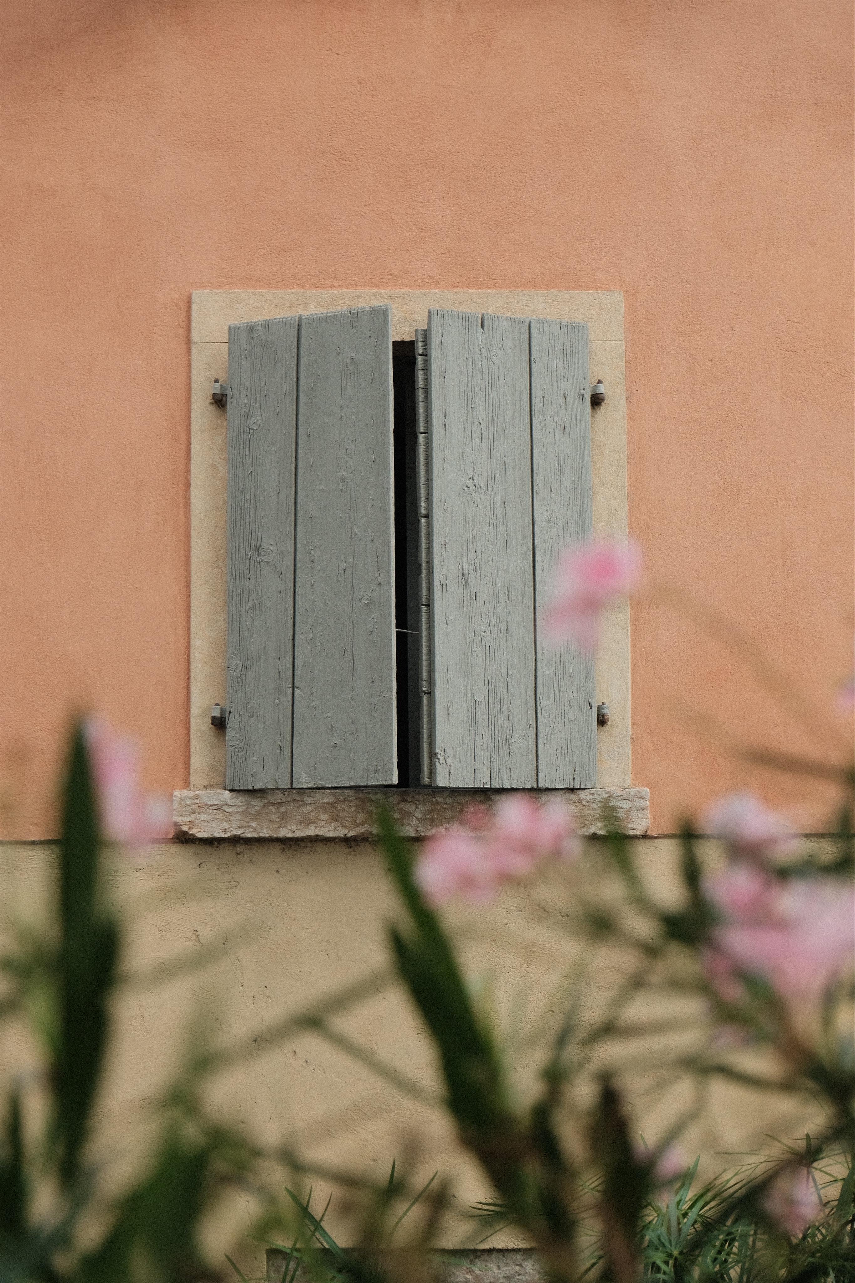 Closed sage / gray wooden shutters on a small window set in a peach stucco wall, with blurred pink flowers and green leaves in the foreground.