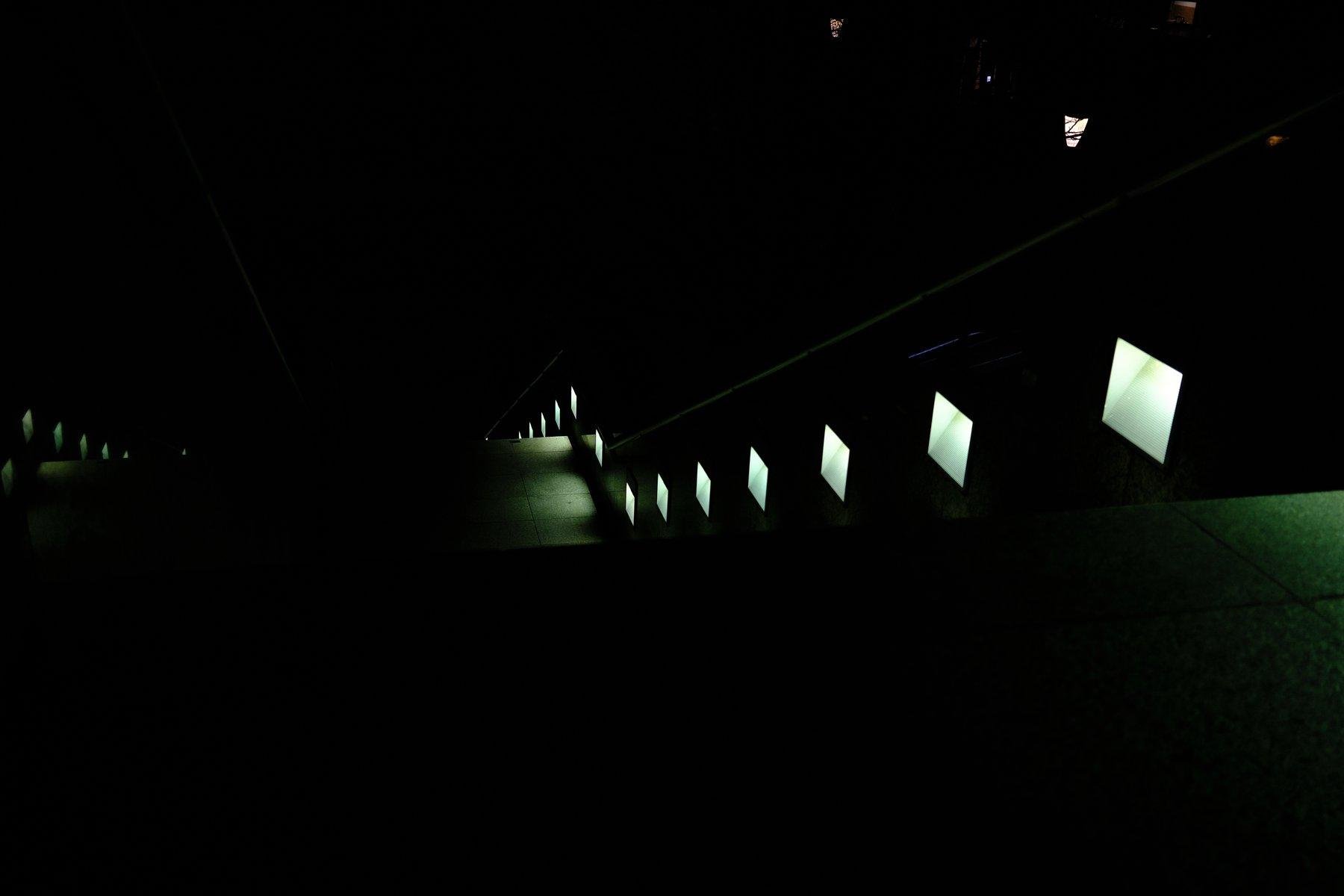 Photo format paysage, de nuit. on voit principalement des carrés de lumières de teinte verdâtre, qui suivent un escalier descendant qu’on voit à peine. On voit aussi à mi-chemin, un palier avec des dalles carrées