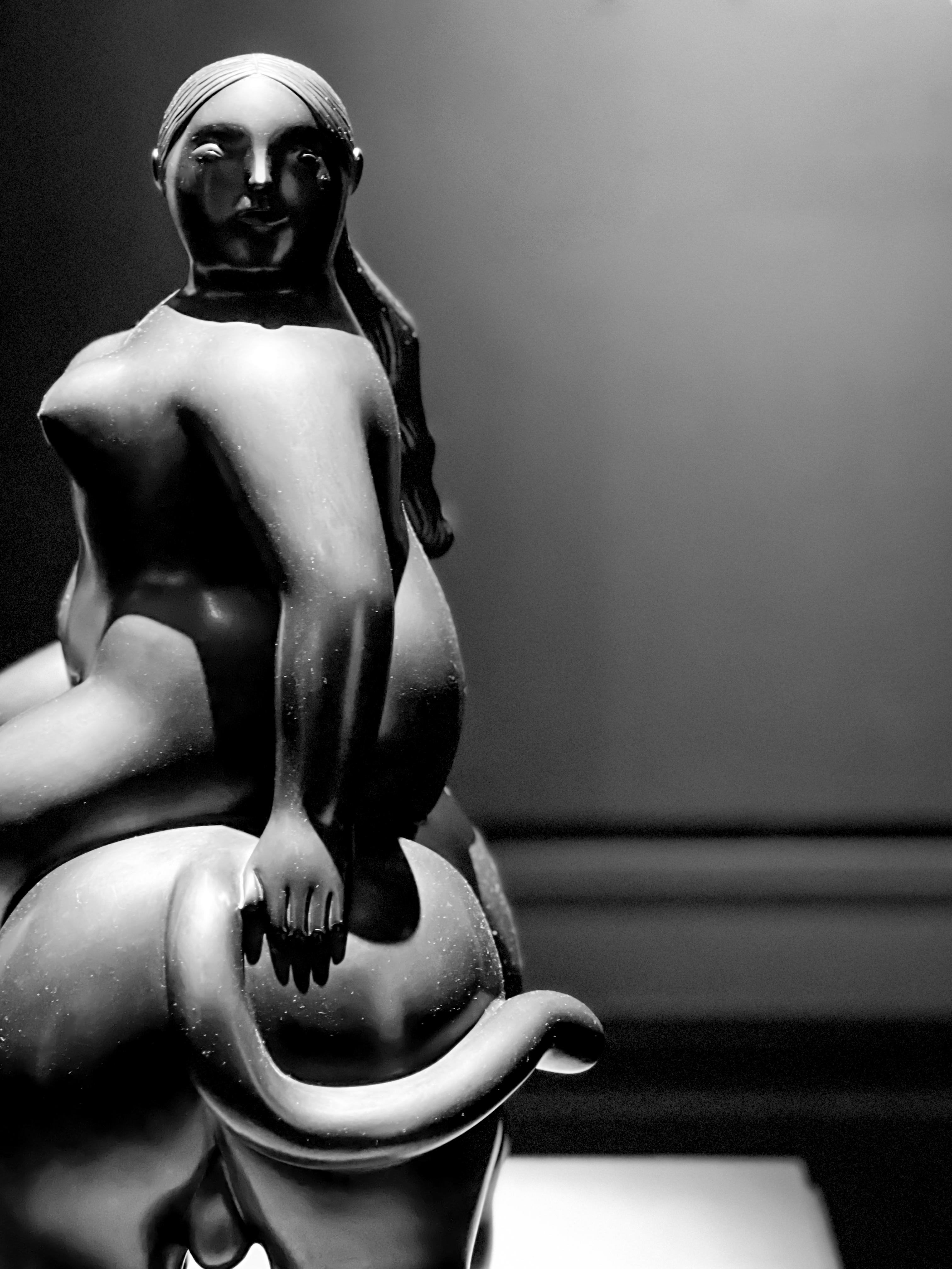 Scultura in bianco e nero di una figura femminile seduta, con un'espressione serena e gli occhi chiusi. La figura è formosa e sensuale, con dettagli anatomici ben definiti. La scultura è realizzata in uno stile classico, con linee morbide e armoniose. Lo sfondo è scuro e uniforme, per focalizzare l'attenzione sulla figura.