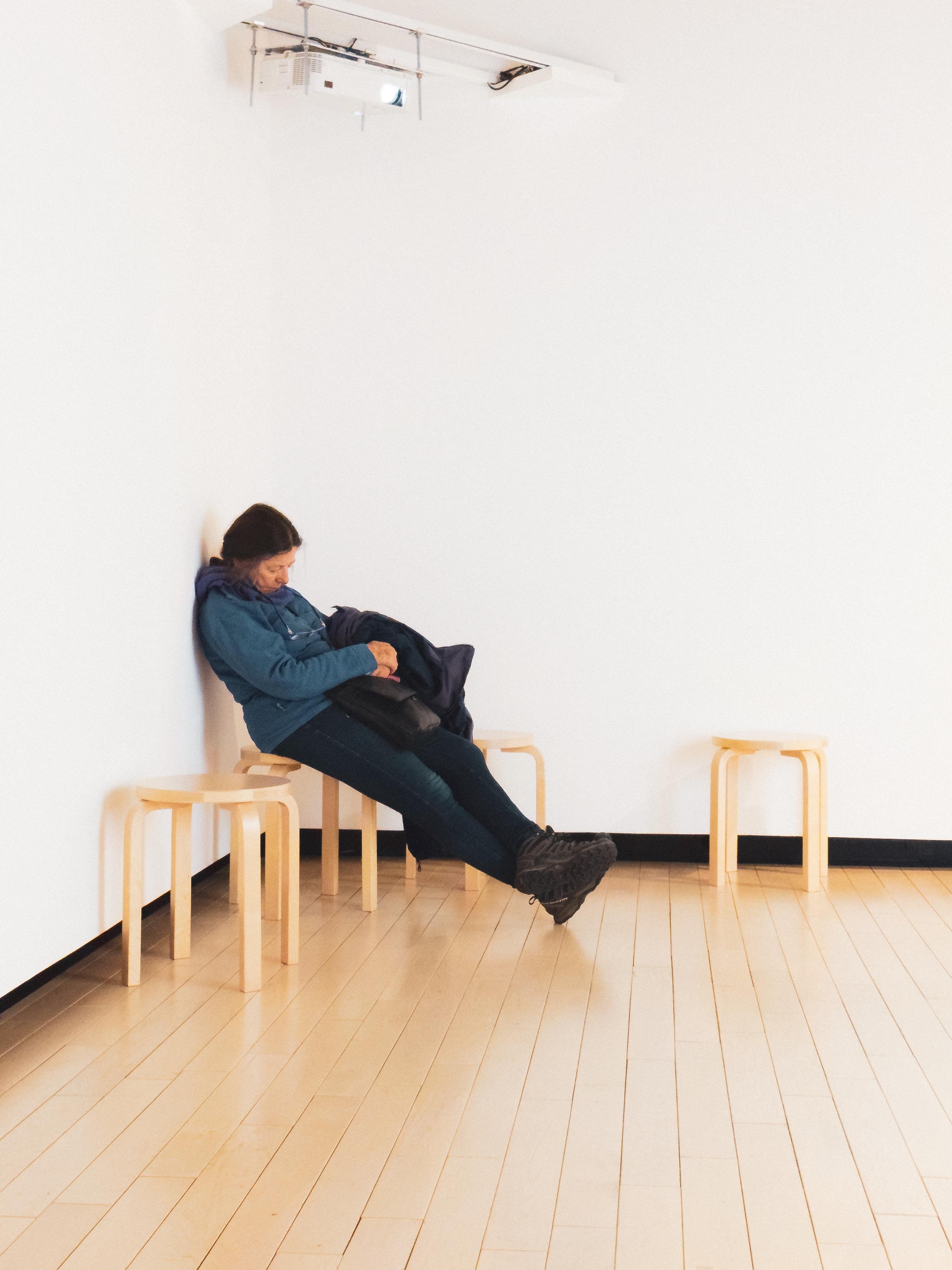 The image depicts a person sitting in a seemingly impossible position, appearing to lean against a wall with their legs outstretched and elevated, above the floor. It creates an optical illusion, likely supported by a hidden structure or effect that makes the position appear unnatural. The setting is a minimalist room with a few stools and a projector on the ceiling.