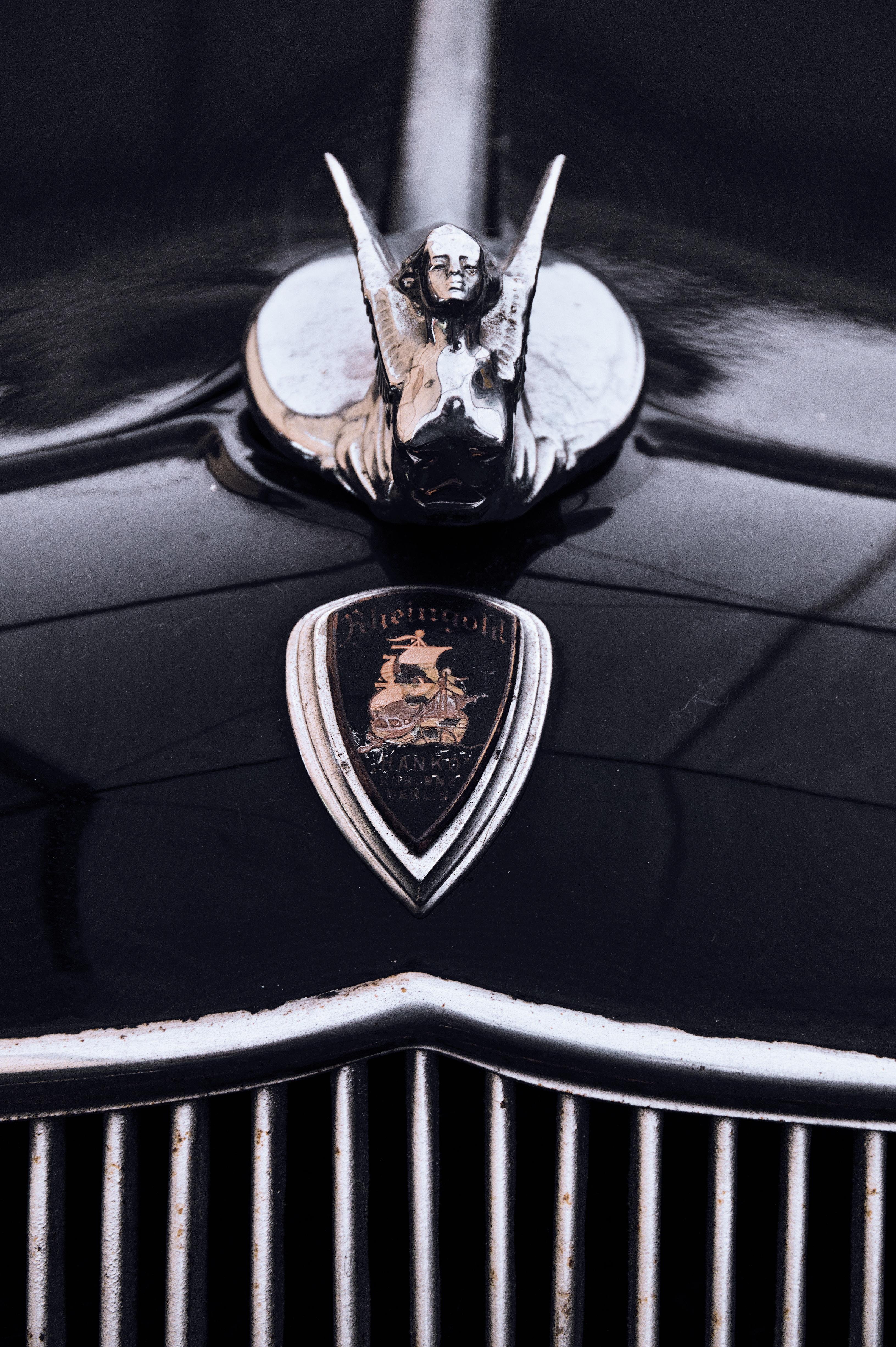 Close-up of a car hood featuring a chrome hood ornament shaped like a figure with wings, positioned above a shield-shaped badge. The badge displays text and an image of a ship, above a chrome grille with vertical bars. The car surface is glossy black.
###
Nahaufnahme einer Motorhaube mit einer verchromten Motorhaubenverzierung in Form einer Figur mit Flügeln, die über einem schildförmigen Abzeichen positioniert ist. Das Abzeichen zeigt den Text und das Bild eines Schiffes über einem verchromten Kühlergrill mit vertikalen Balken. Die Oberfläche des Autos ist glänzend schwarz.