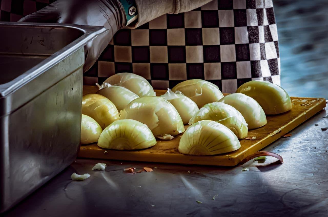 The image shows a person wearing a checkered apron or uniform, cutting onions. There are several halved onions on a cutting board, and a container is visible nearby. The scene seems to be in a kitchen or food preparation area.