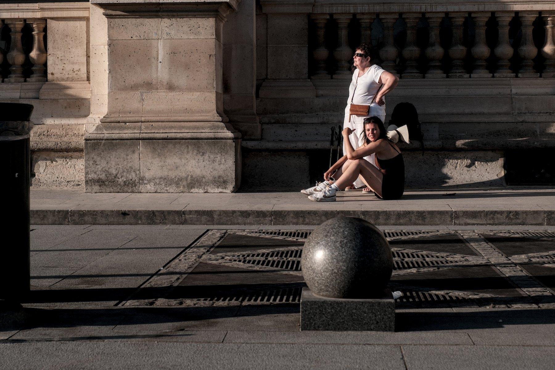 Photo horizontale. Un femme debout toute habillée de blanc, observe l’éclipse avec les protections adéquate, vers la gauche de l’image. À sa gauche, devant elle de notre point de vue, une jeune femme en short et débardeur noirs, assise par terre, tourne la tête vers la caméra en souriant