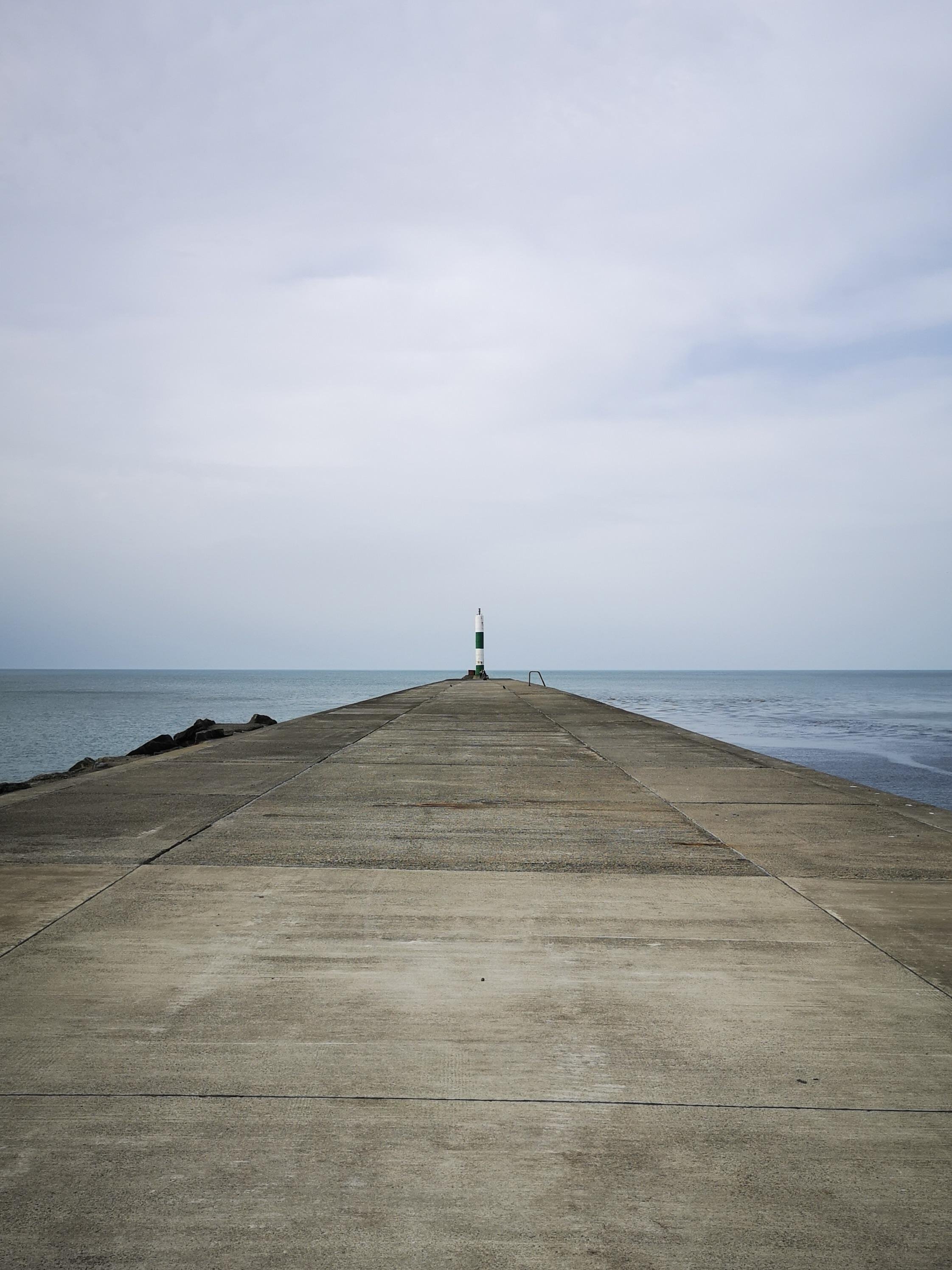 Long concrete pier extending into a calm sea under an overcast sky, with a small green-and-white navigation marker at the far end and rocks along the left side.