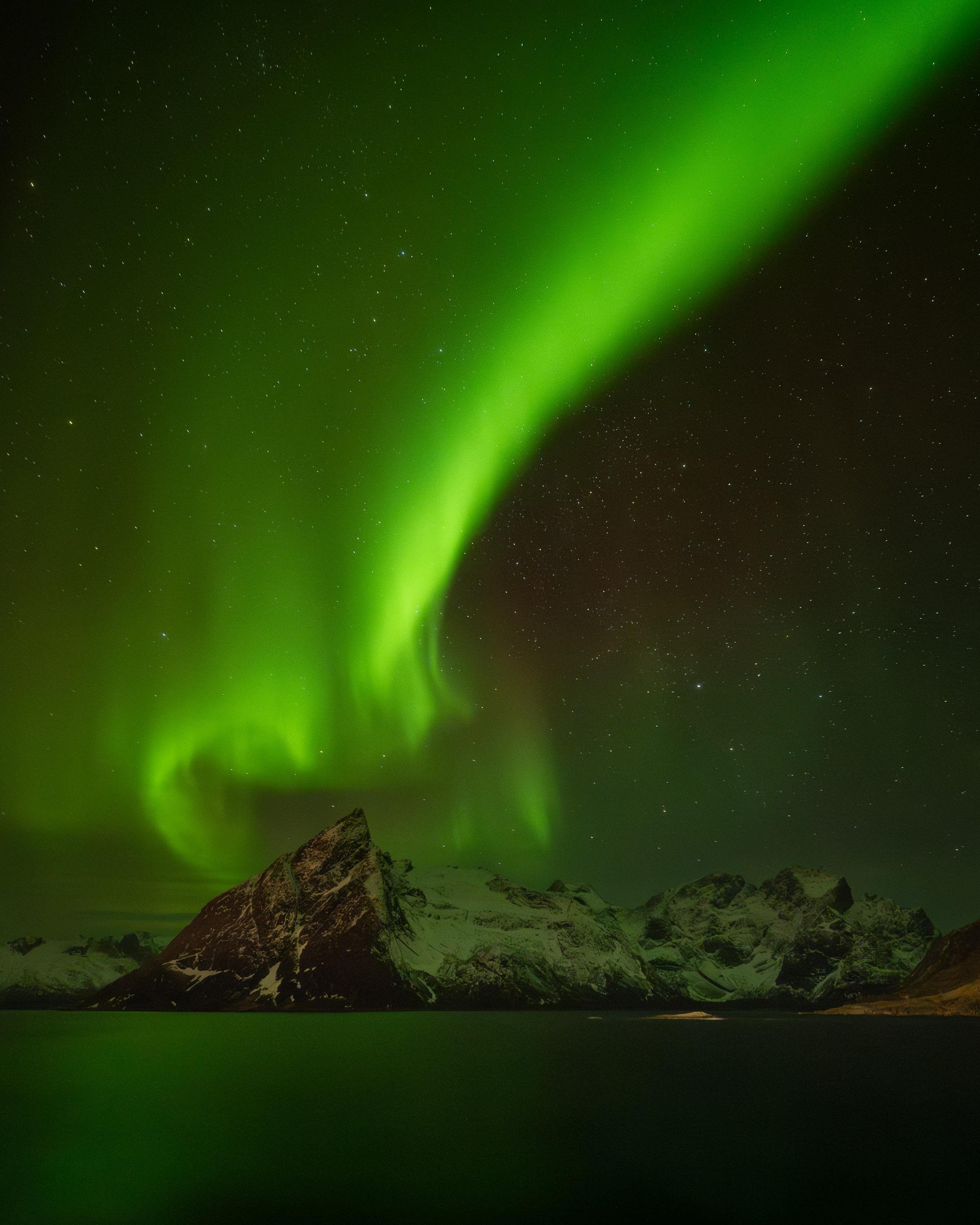 This image shows the Northern Lights, or Aurora Borealis, above a snowy mountainous landscape. The sky is illuminated with vibrant green lights against a backdrop of stars. The scene appears to be a nighttime view capturing this natural light display.