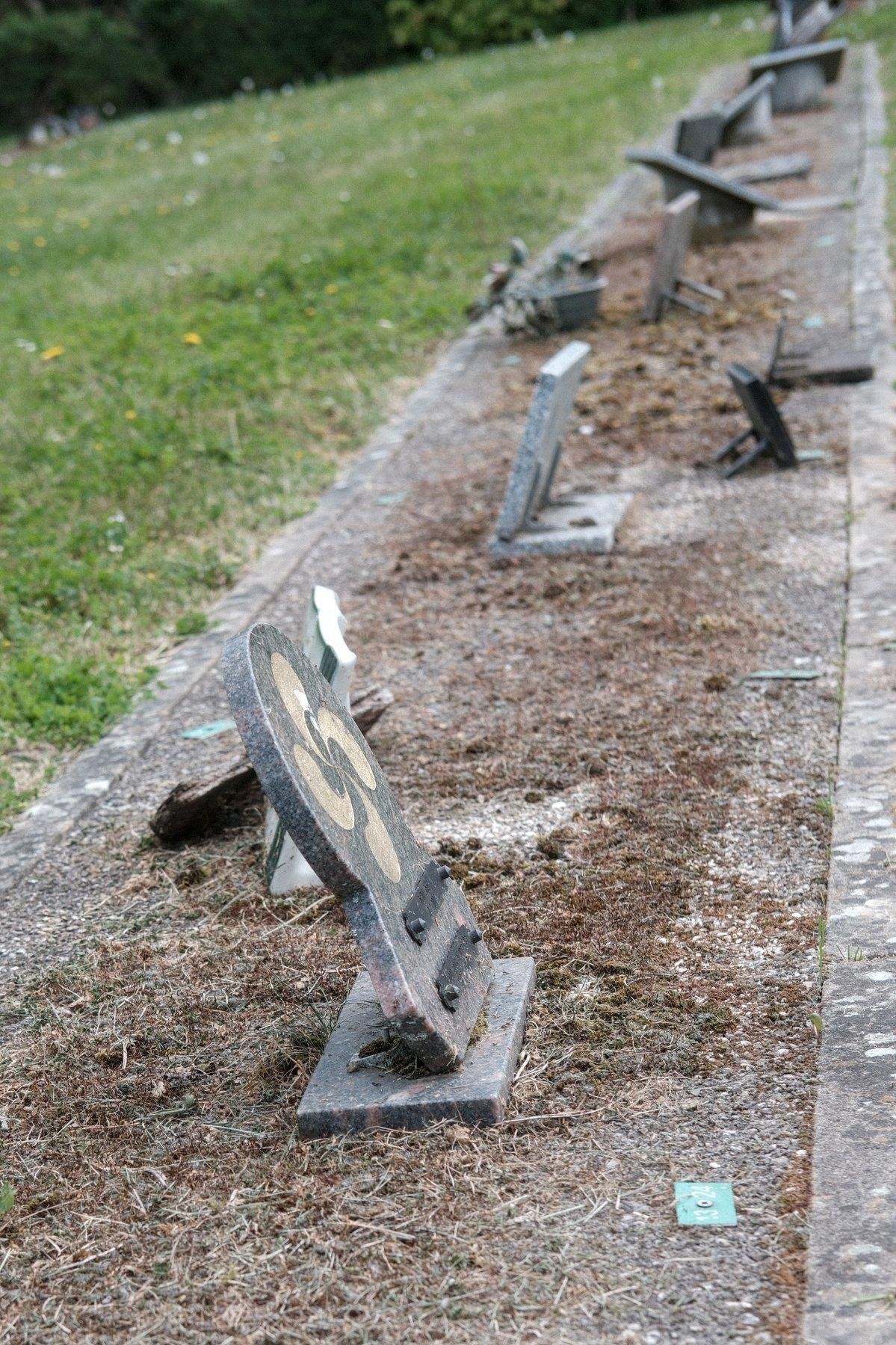 Photo verticale. Sur une bande de béton sont posées à intervals régulier des plaque funéraires de petites tailles. En dehors de la bande de béton, sur la gauche, de la pelouse
