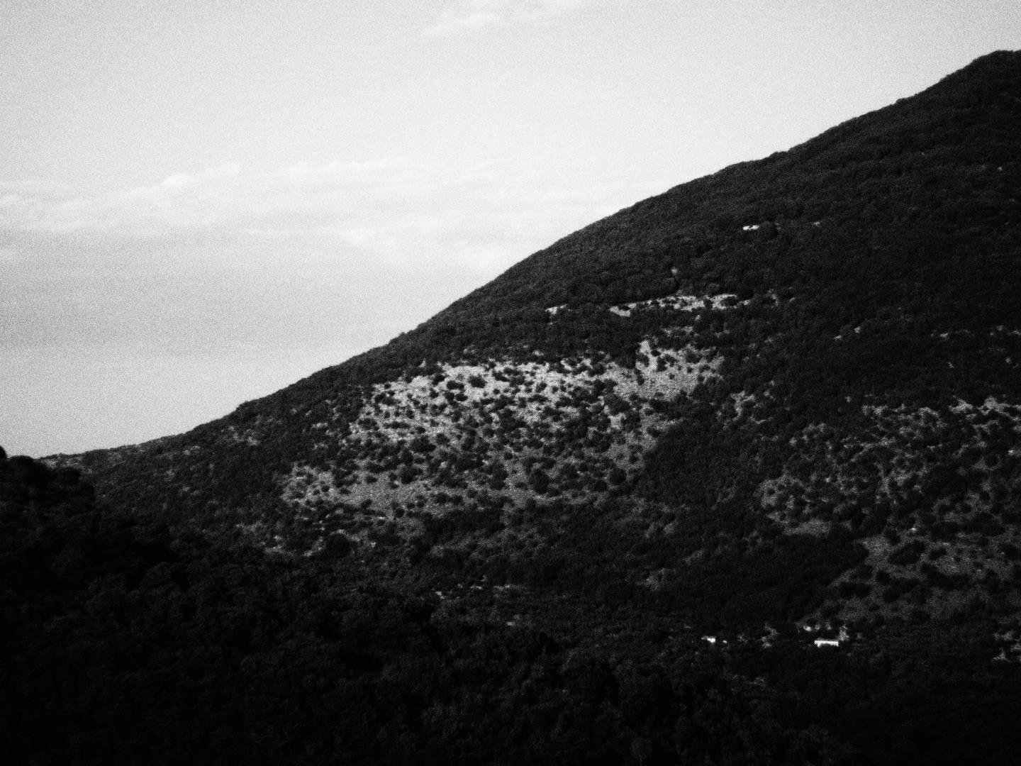 Inquadratura divisa in due dalla doppia curva creata da una montagna ricoperta dalla vegetazione, che si staglia contro il cielo del pomeriggio estivo.