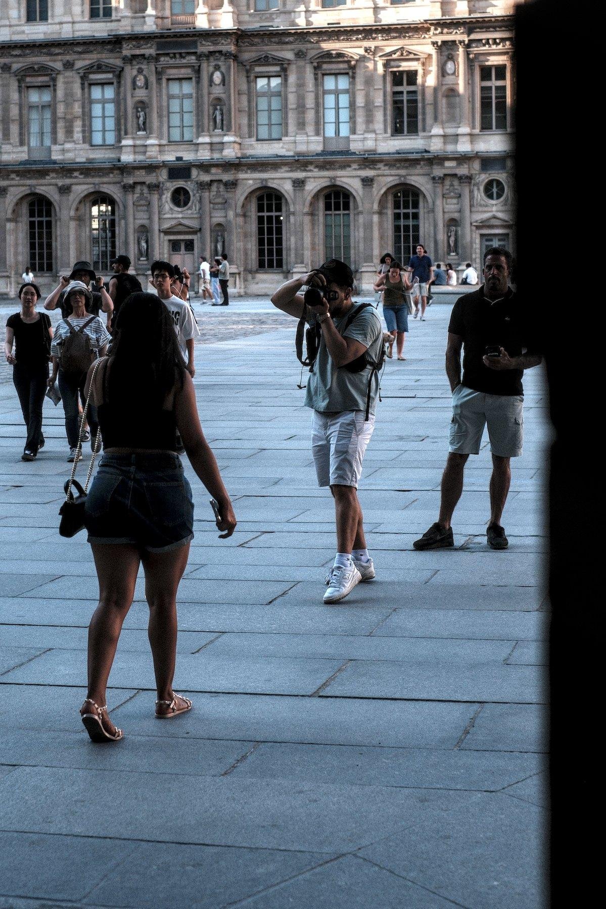 Photo verticale. Une jeune femme de dos marche vers un homme qui la prends en photo sur une place piétonne