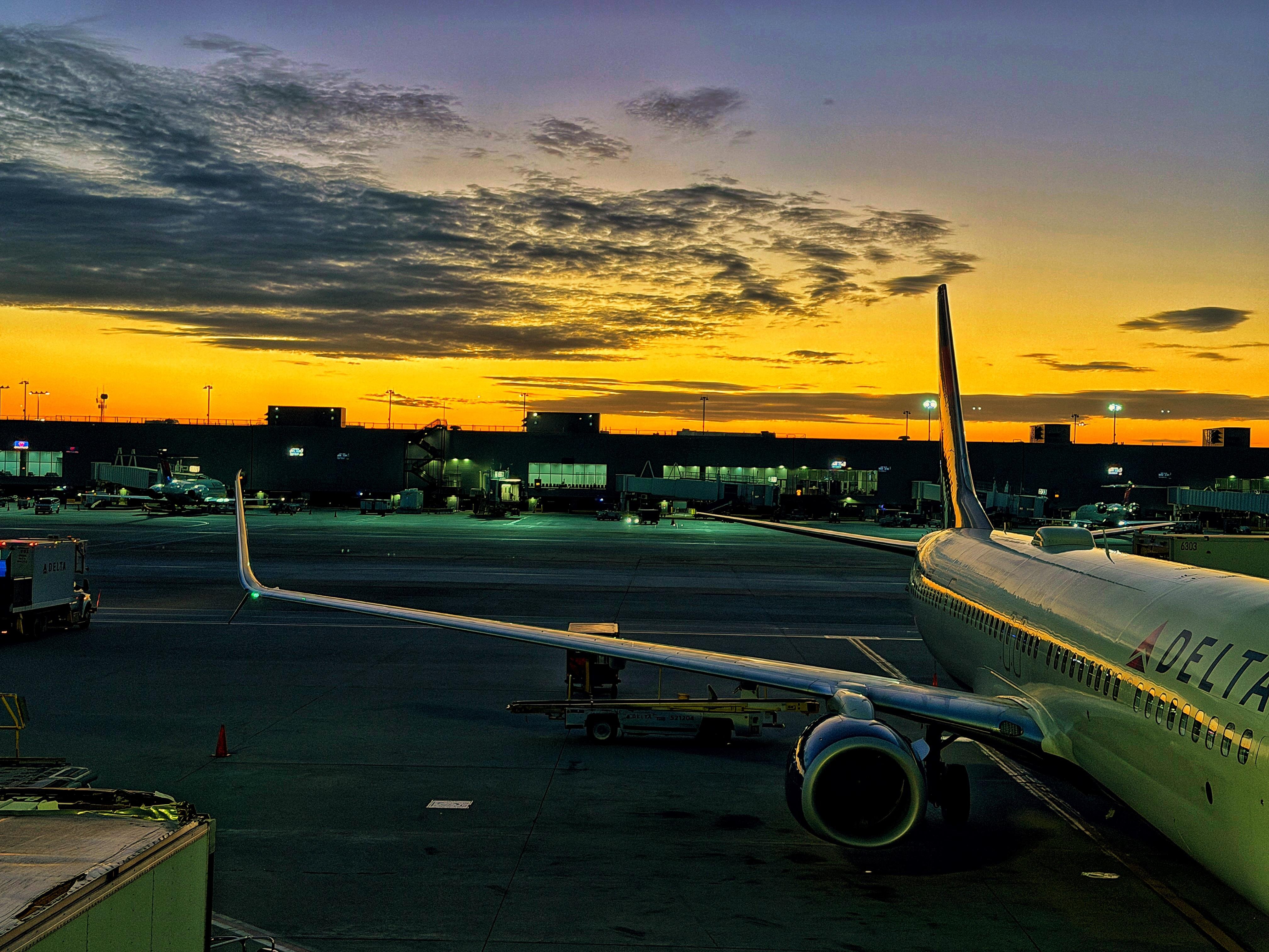 Passenger airplane parked at an airport gate during sunset, with dark clouds and an orange sky in the background. The airline's name is partially visible on the fuselage. Illuminated airport terminal buildings and equipment are in the distance, with ground service vehicles near the aircraft.