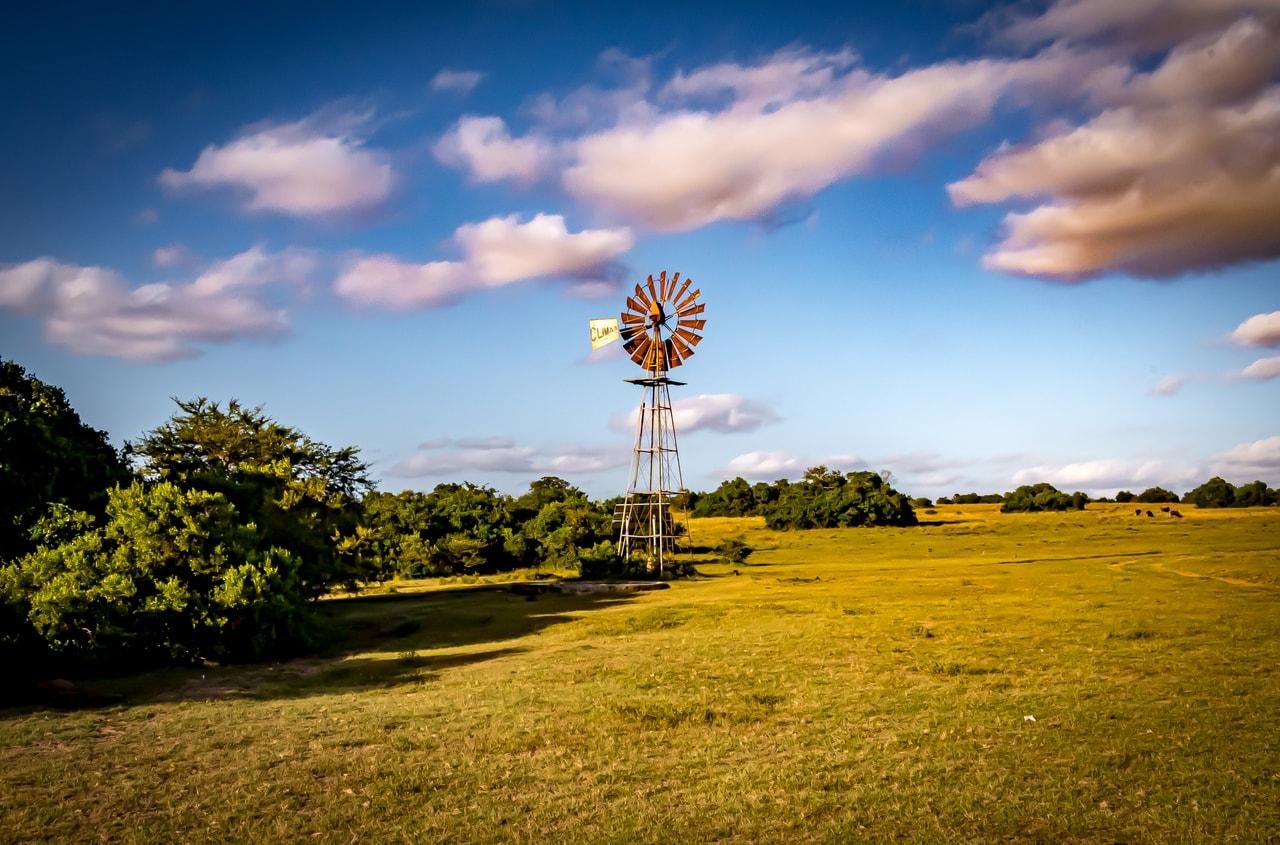 The image shows a rural landscape with a windmill on a grassy field. The sky is blue with scattered clouds, and there are some trees around the field. The scene has a peaceful and open countryside feel.