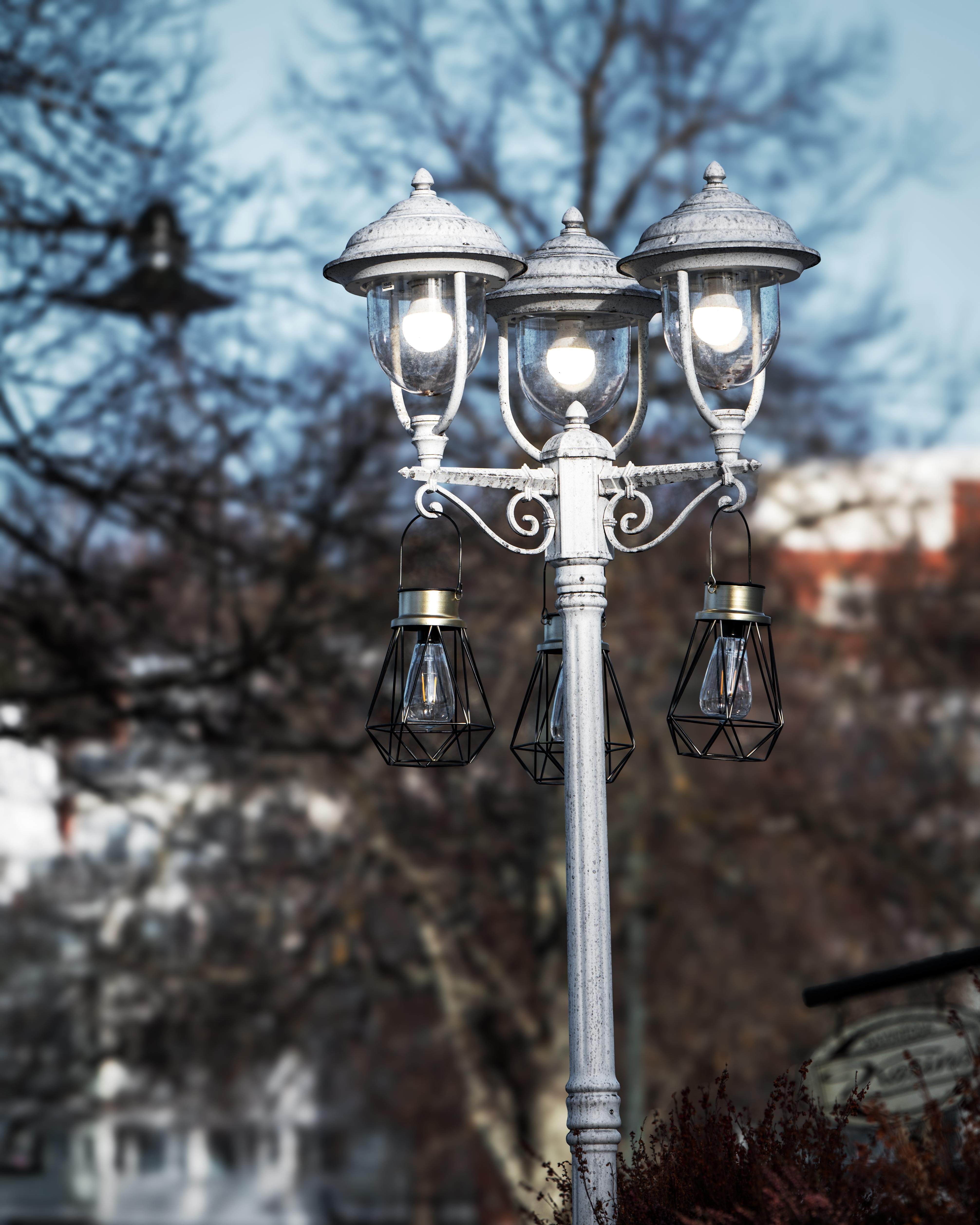 The image shows a decorative street lamp with three illuminated bulbs, each housed in a glass enclosure with a dome-shaped cover. Below the main lamps, there are three additional hanging lantern-style lights. The lamp post is set against a blurred background of trees and buildings, suggesting an outdoor setting, possibly in a park or street area.