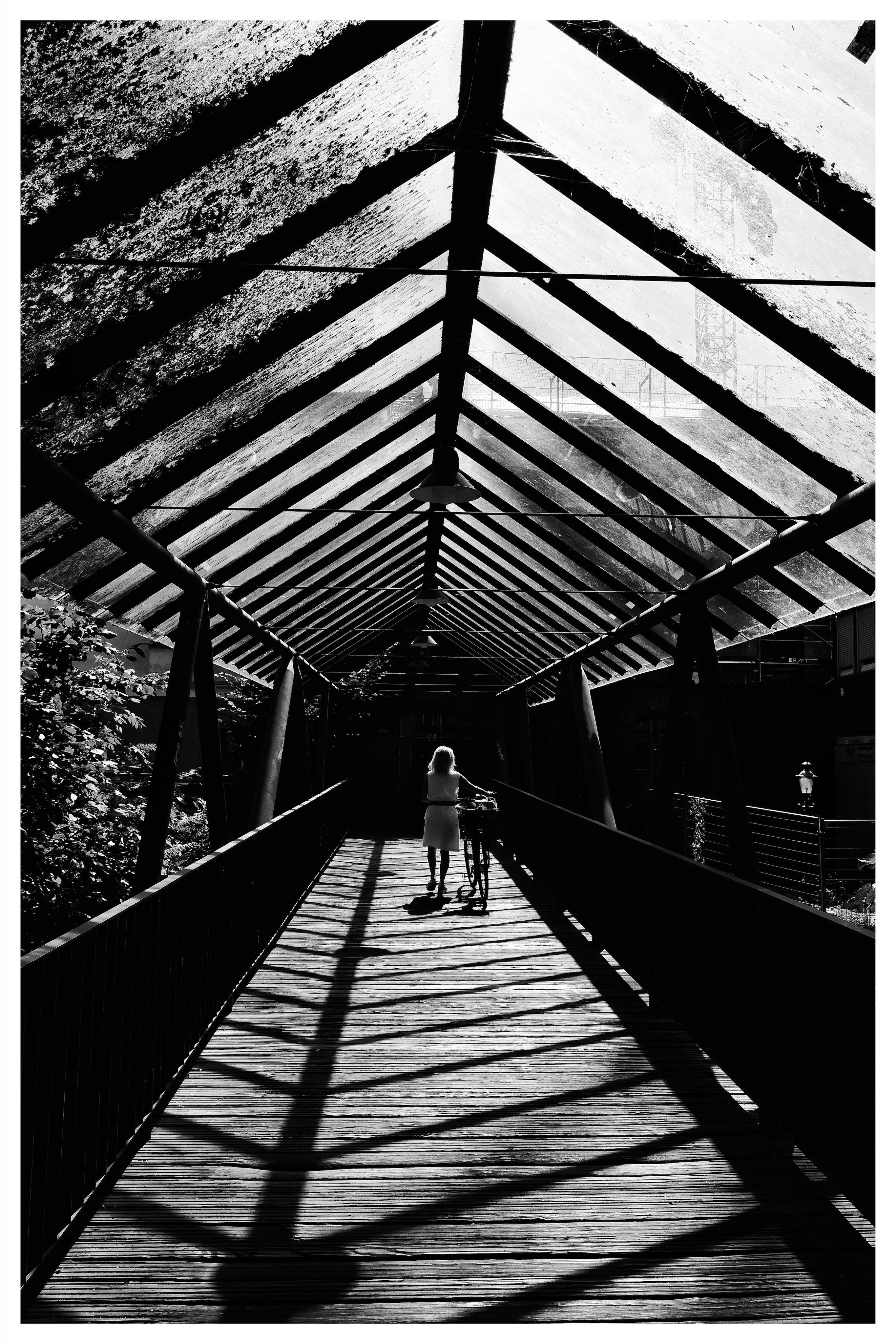 Black-and-white photo of a covered pedestrian bridge with exposed roof beams forming a repeating triangular pattern; a person walks away from the camera pushing a bicycle along the wooden walkway, with strong beam shadows stretching across the floor.