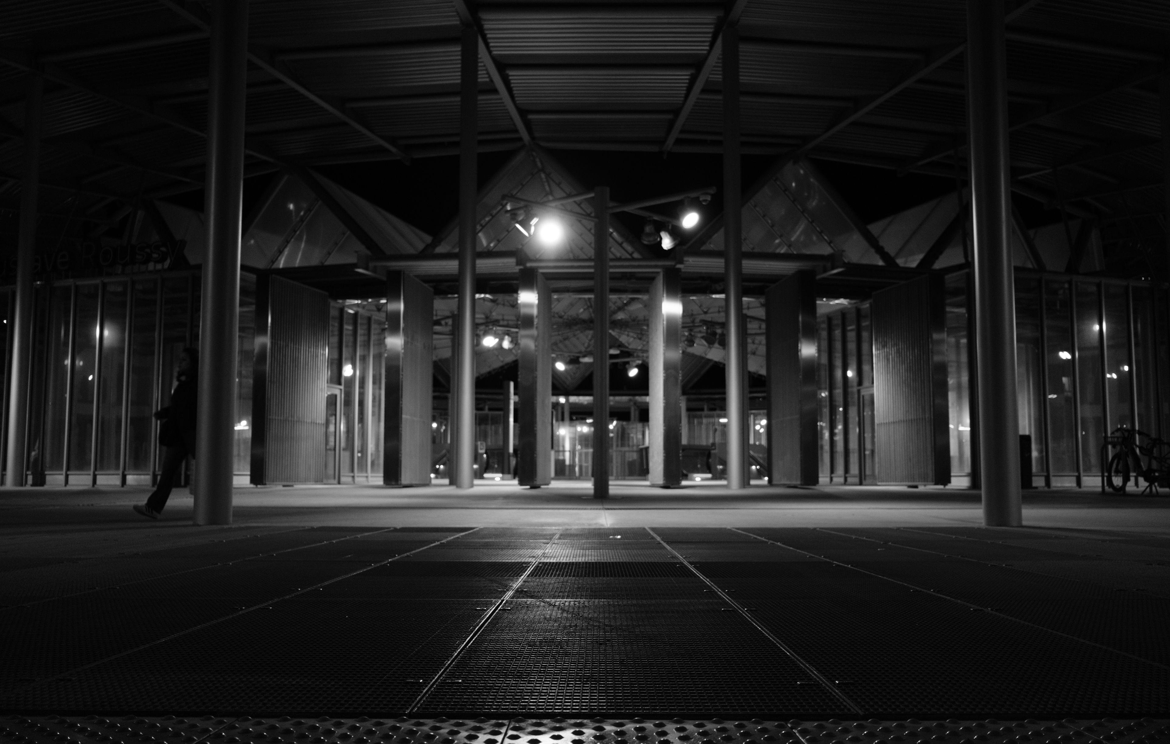 Photo format paysage en noir et blanc, de nuit, avec au premier plan des grilles au sol et un bout de picots pour malvoyants avant la chaussée,  et plus flou, des poteaux et un toit formant une entrée de la station Villejuif Gustave Roussy, dont de la lumière émane