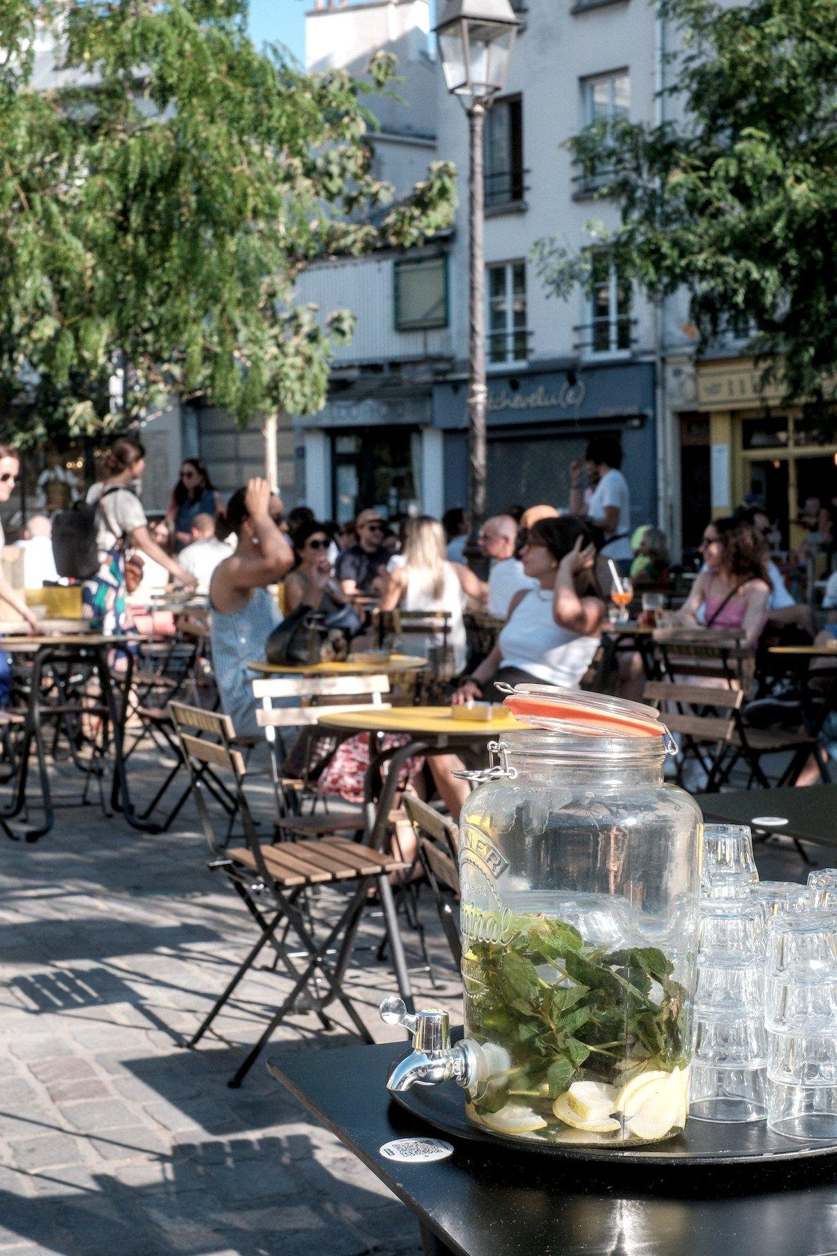 Photo verticale. Sur une terrasse estivale installée sur une place pavée, peu de tables libres. Au premier plan, posée sur une table noire et ronde, une fontaine en verre (grosse bonbonne avec couvercle à joint orange et un petit robinet tout en bas) avec de l’eau et du citron et de la menthe infusant dedans, avec des verres empilés à l’envers à côté