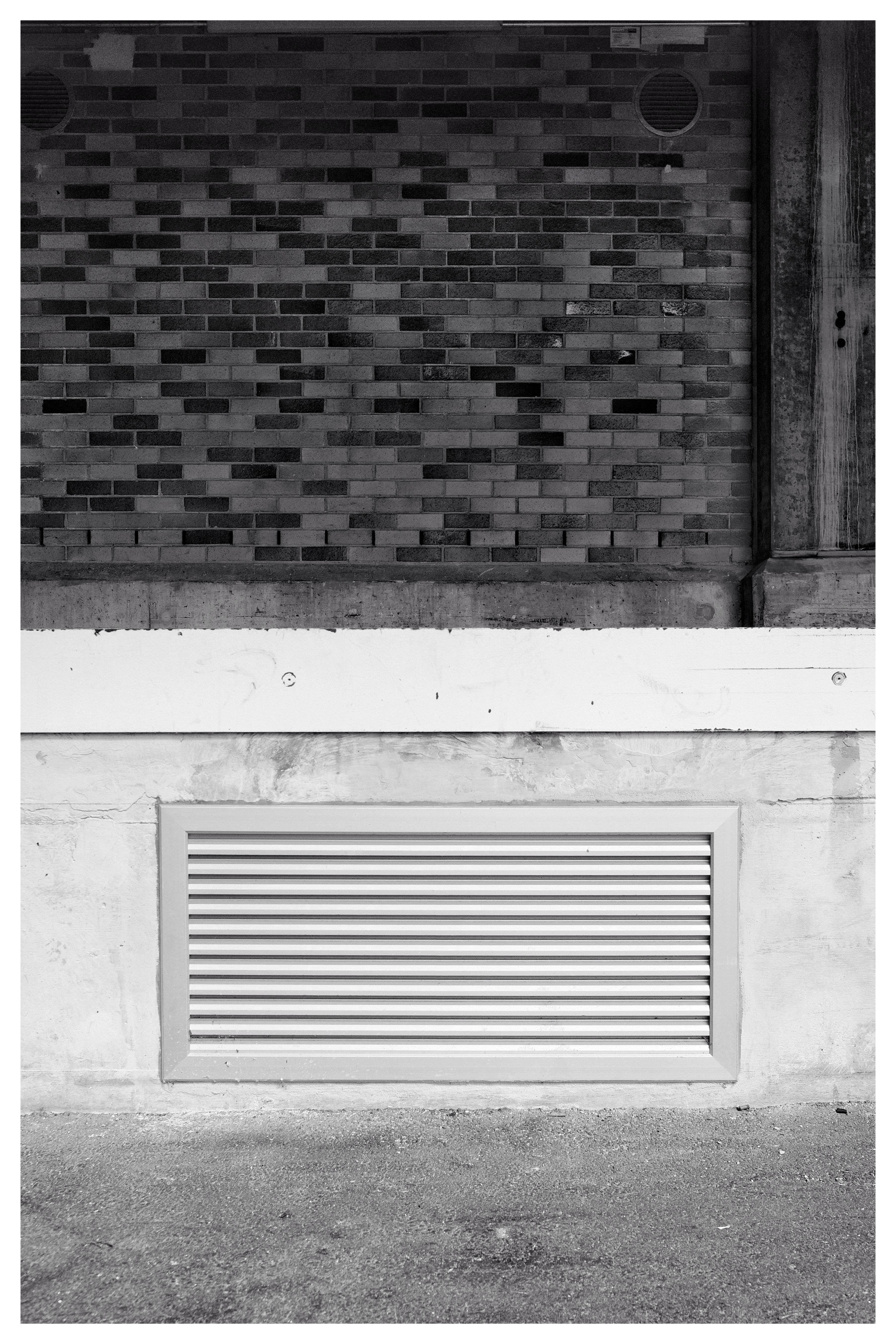Black and white image depicting a building exterior. The top half features a brick wall with different shades and a round vent in the upper right corner. Below, a concrete section culminates with a rectangular metal vent near the bottom center. The ground surface appears rough and textured.