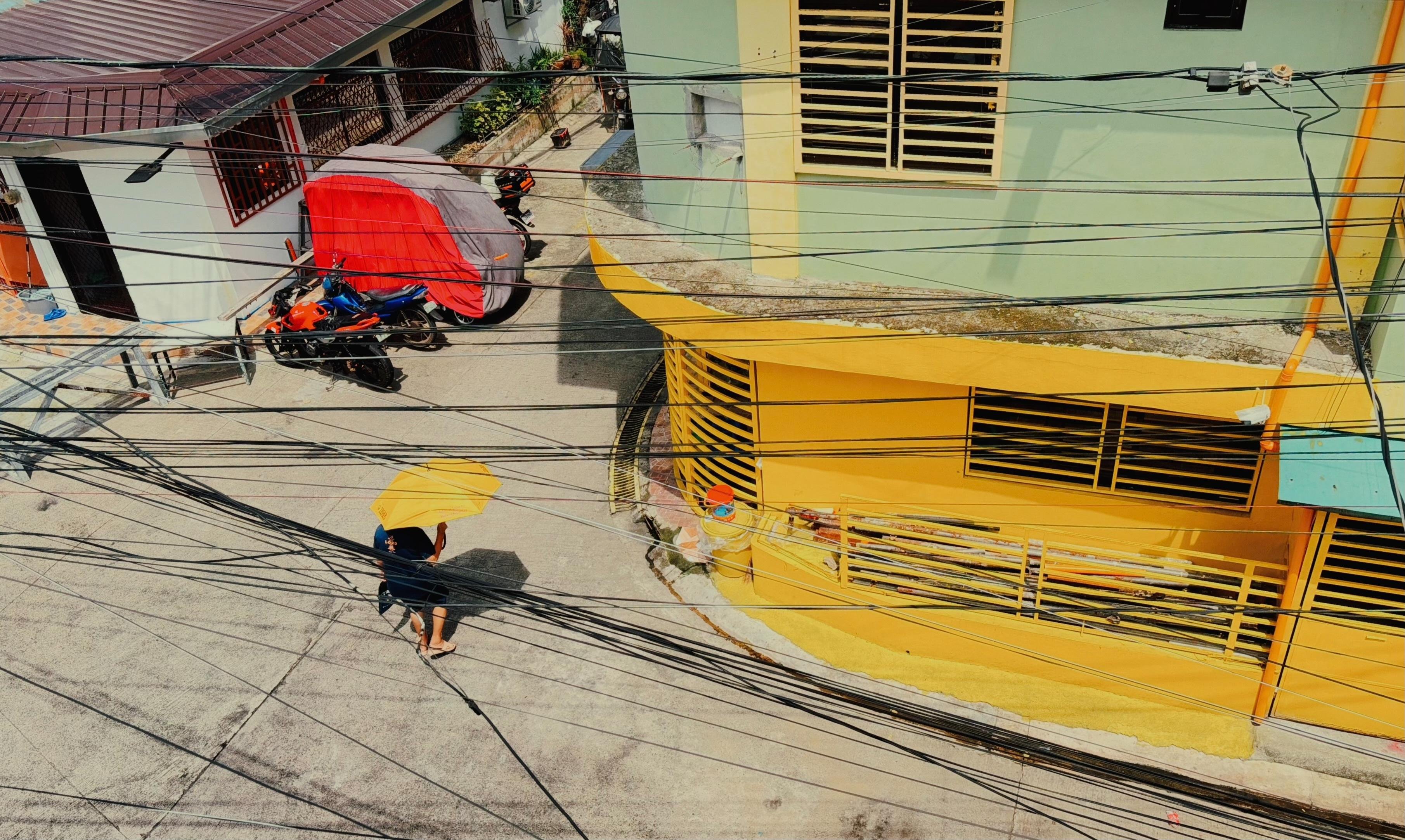 Overhead view of a narrow street corner with a person carrying a yellow umbrella, dense black utility wires crossing the frame, a yellow building with louvered windows on the right, and a motorcycle beside a red canopy near a gray building on the left.
