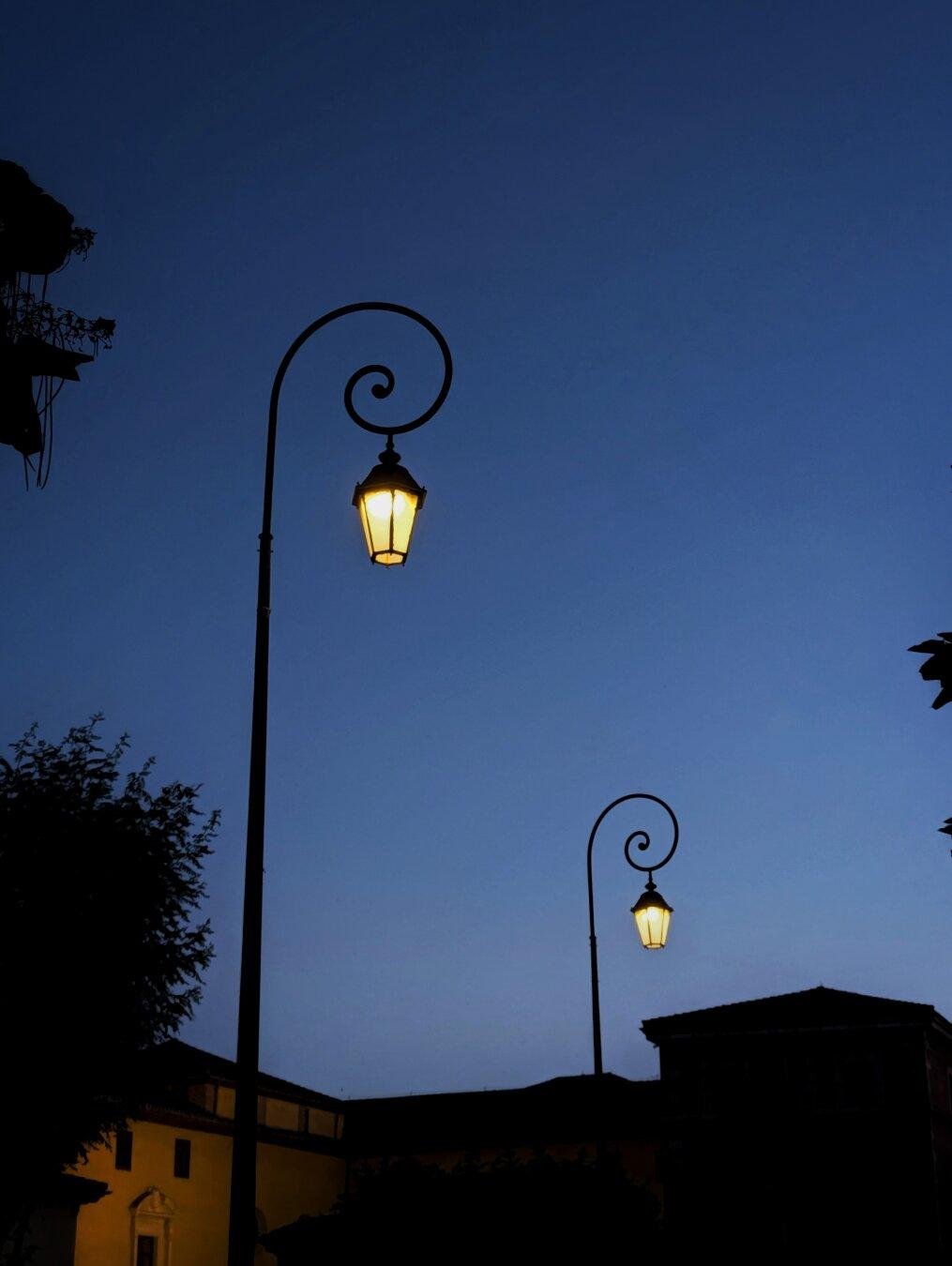En una vista del cielo nocturno, destacan dos farolas altas. Los postes que sujetan cada farol tienen un diseño delicado, como de un zarzillo de una planta que se enrosca sobre sí mismo en una espiral. Los faroles destacan con su luz amarilla en el cielo de un azul oscuro. La imagen transmite mucha calma.