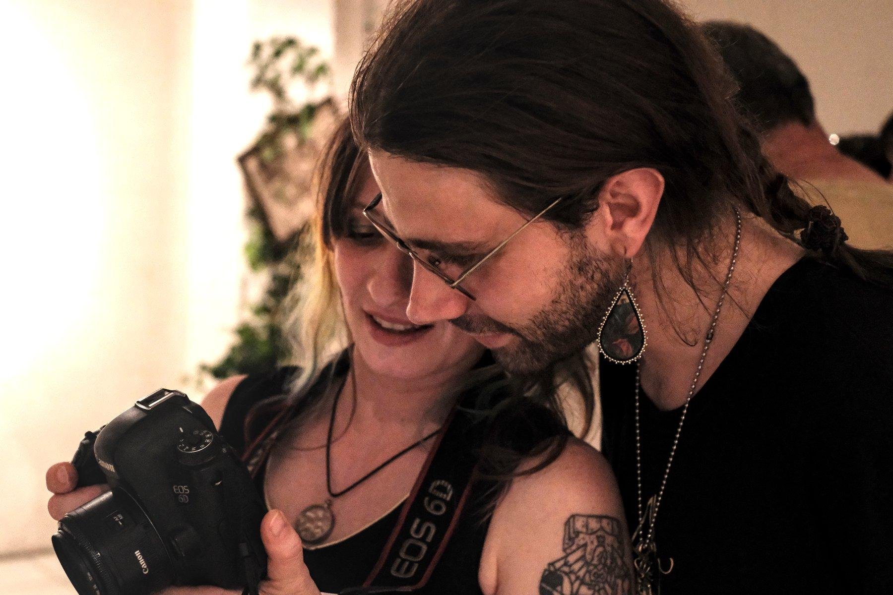 Photo horizontale. Une jeune femme tatouée à l’épaule gauche montre l’écran d’un appareil photo à un homme aux cheveux longs qui porte une boucle d’oreille avec un pendant en forme de goutte assez gros
