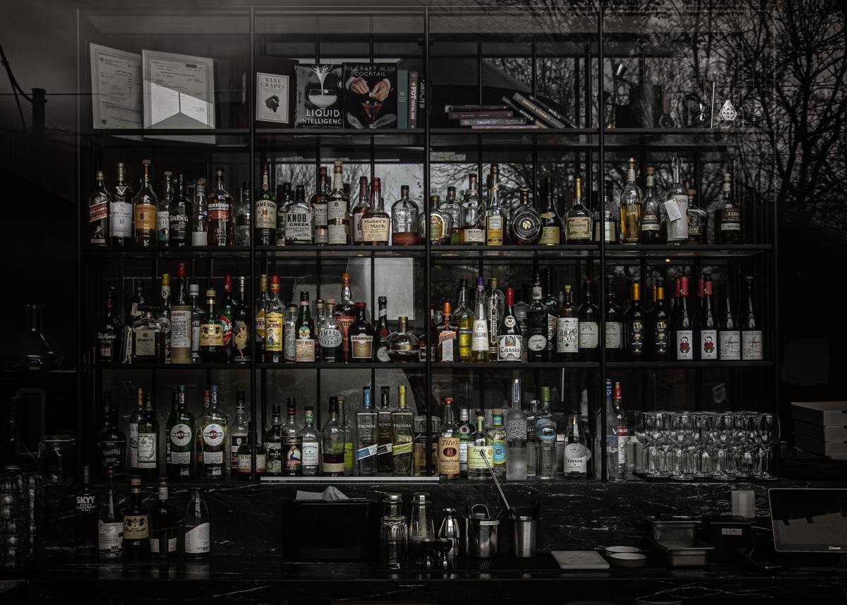 Dimly lit bar shelf filled with numerous bottles of liquor organized across several rows. The shelf is made of dark metal, contrasting with the array of colorful labels on bottles. Glasses are neatly arranged on the lower right side. Above the bottles, books and menus are visible, suggesting a well-stocked and organized bar setting. Reflected tree branches in the background create a subtle, moody atmosphere.