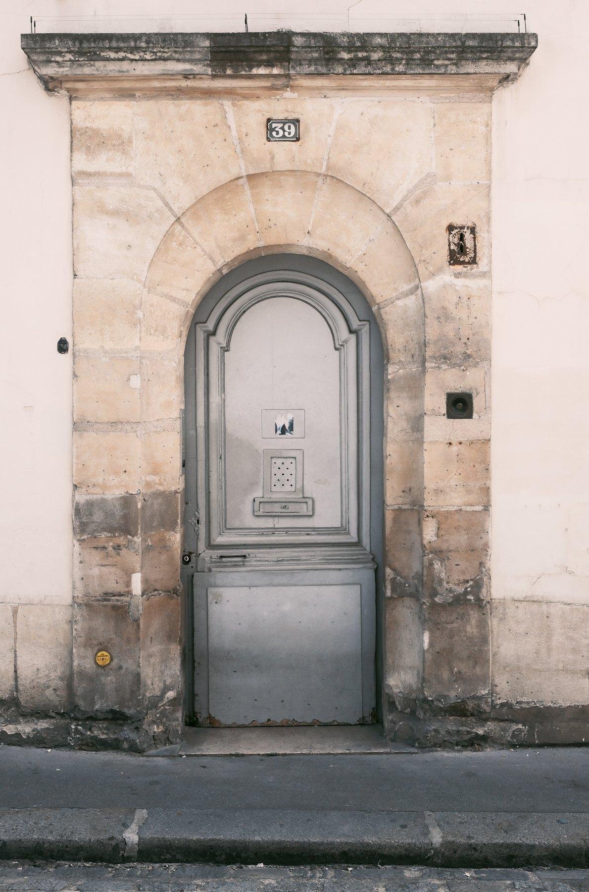 Photo verticale. Une porte dans un cadre en pierre qui dessine une arche, de tout dans les teintes beiges, sauf la porte, grise