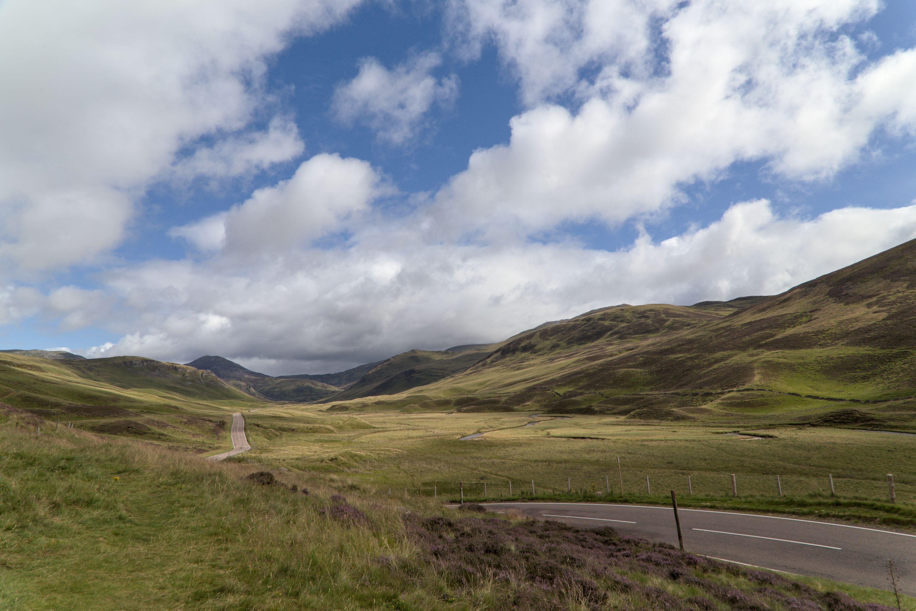 Photo horizontale. Les Highlands écossais et ses colinnes verdoyante. Une route les traverses, partant du premier plan en bas à droite vers l’horizon.
