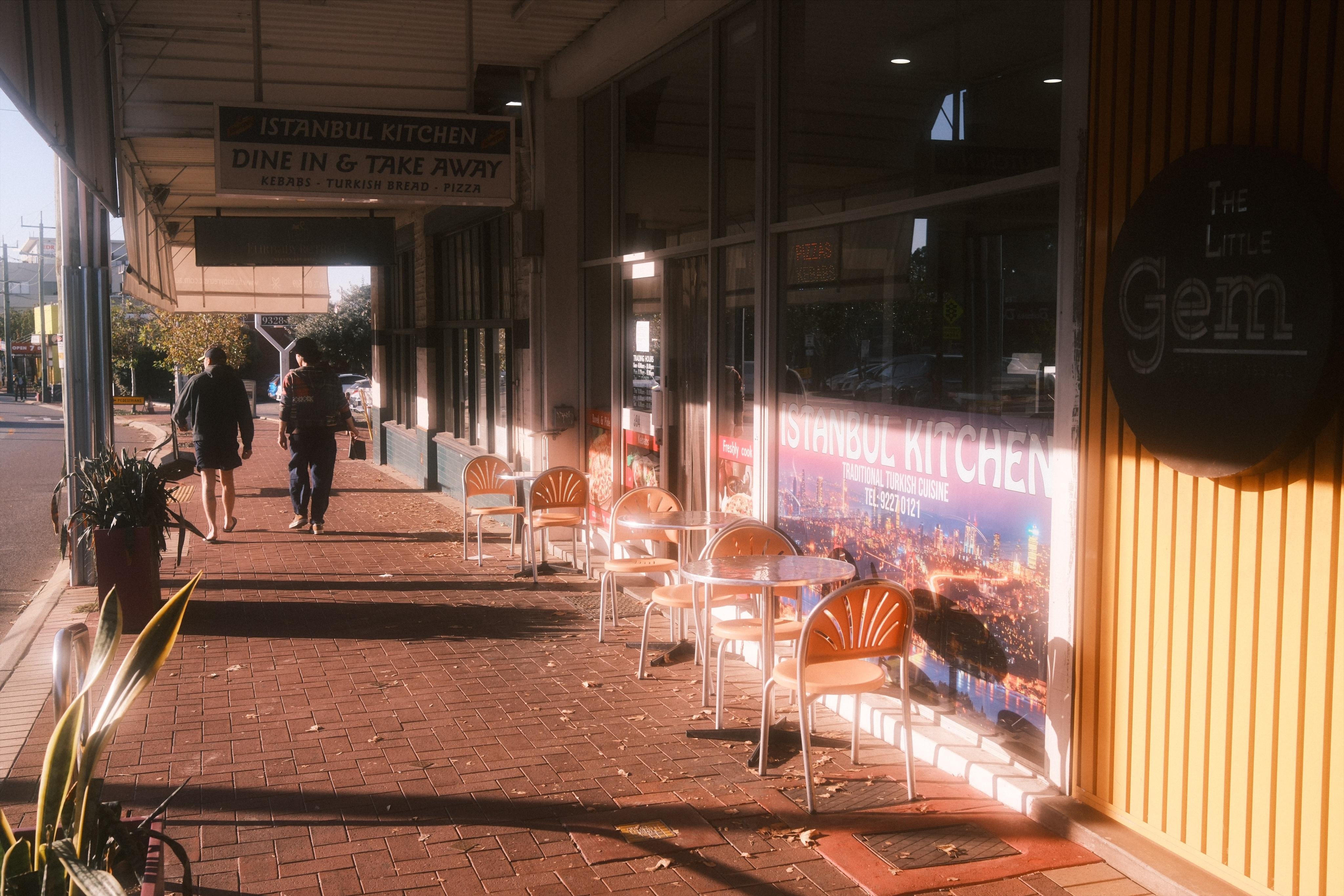 Covered sidewalk with brick paving beside storefront windows for “Istanbul Kitchen,” with several small outdoor tables and chairs lined up along the wall; two pedestrians walk away in the distance, and a round sign reading “The Little Gem” is mounted on the right.
