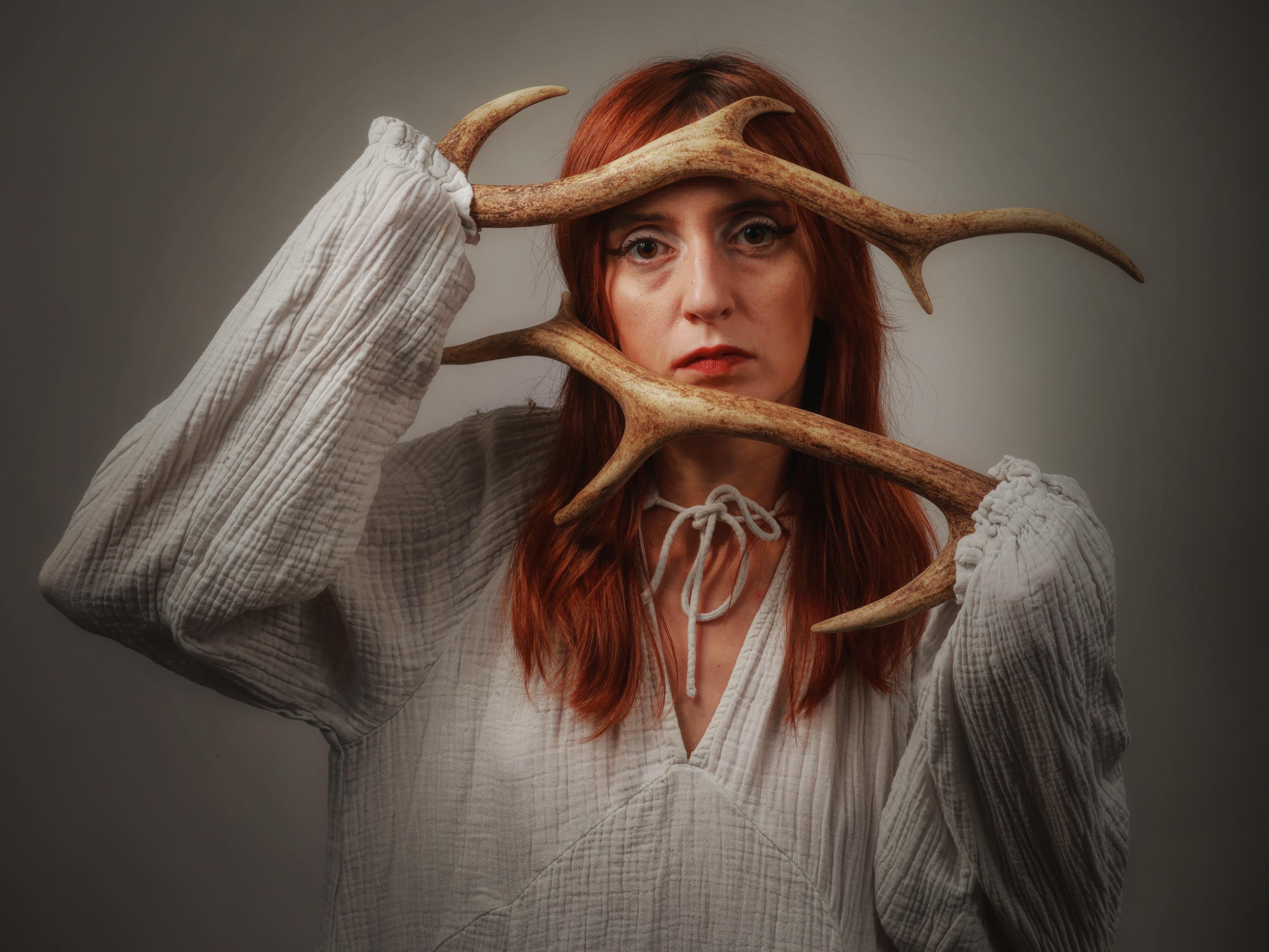 Woman with long red hair holding two antlers across her face, wearing a white, textured, long-sleeved blouse. The background is a neutral gray, and she has a contemplative expression.