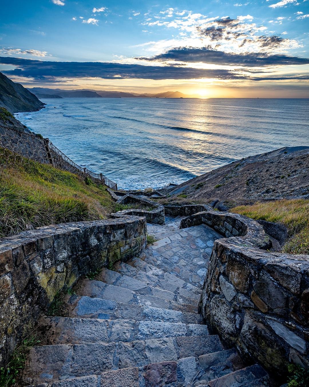 Stone steps bordered by moss-covered stone walls leading down a grassy hillside towards a rocky coastline and expansive ocean. The sun is setting on the horizon, casting orange and yellow hues across the sky and reflecting on the water. Layered clouds with a mix of blue sky create a dramatic backdrop.