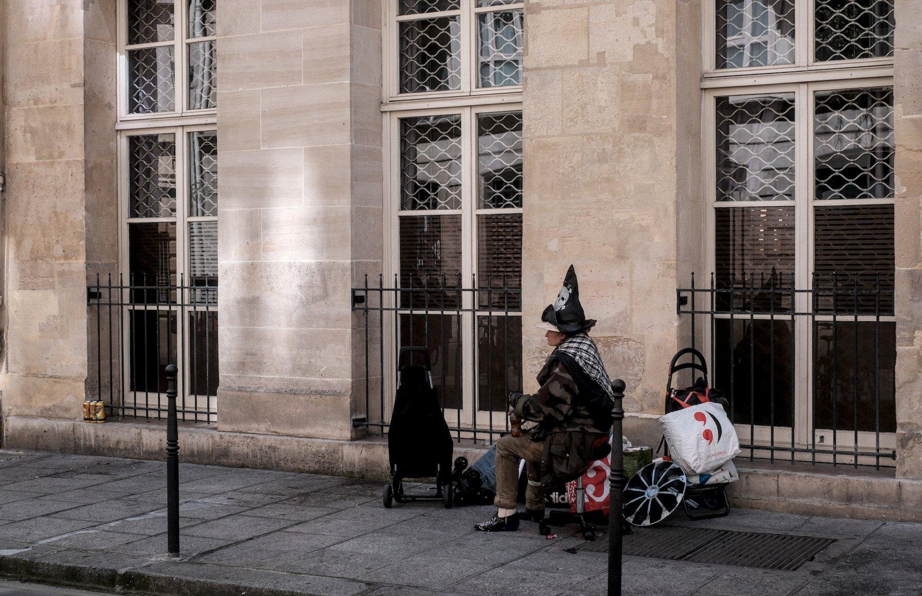 Photo horizontale. Sur un trottoir, une dame est assise sur une chaise de bureau sans dossier, avec deux caddie et des sacs pendus dessus, une jante de voiture, qui sont probablement toutes ses possessions… Elle porte un chapeau pointu noir de sorcière avec une tête de mort de pirate imprimée dessus