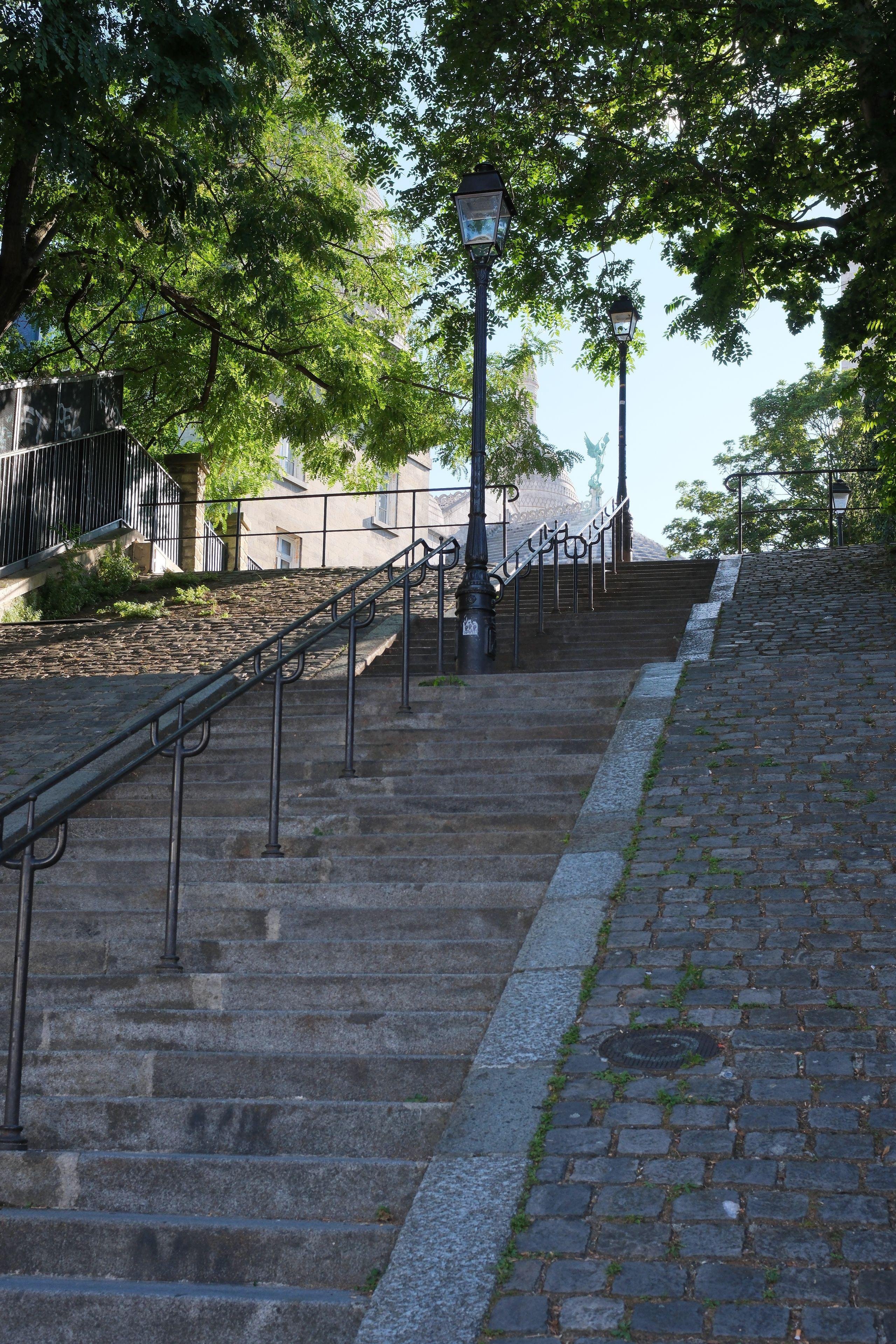 Stone stairs flanked by cobblestone paths and black metal handrails, with street lamps and leafy green trees overhead during daytime. We can see a statue of an angel at the top