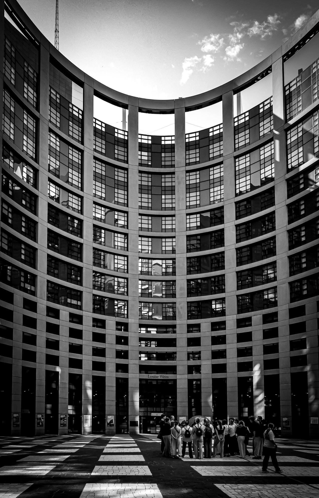 Curved, high-rise building facade with multiple rows of reflective windows. The foreground features a black and white checkered pavement. A group of people stands near the entrance labeled "Louise Weiss," casting shadows on the ground. The sky above is partly cloudy.