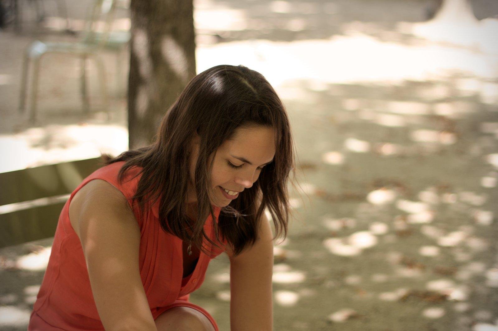 Photo format portrait d’une fille brune, cheveux longs, assise sur un banc sous l’ombre des arbres, souriante, regardant vers ses pieds hors cadre