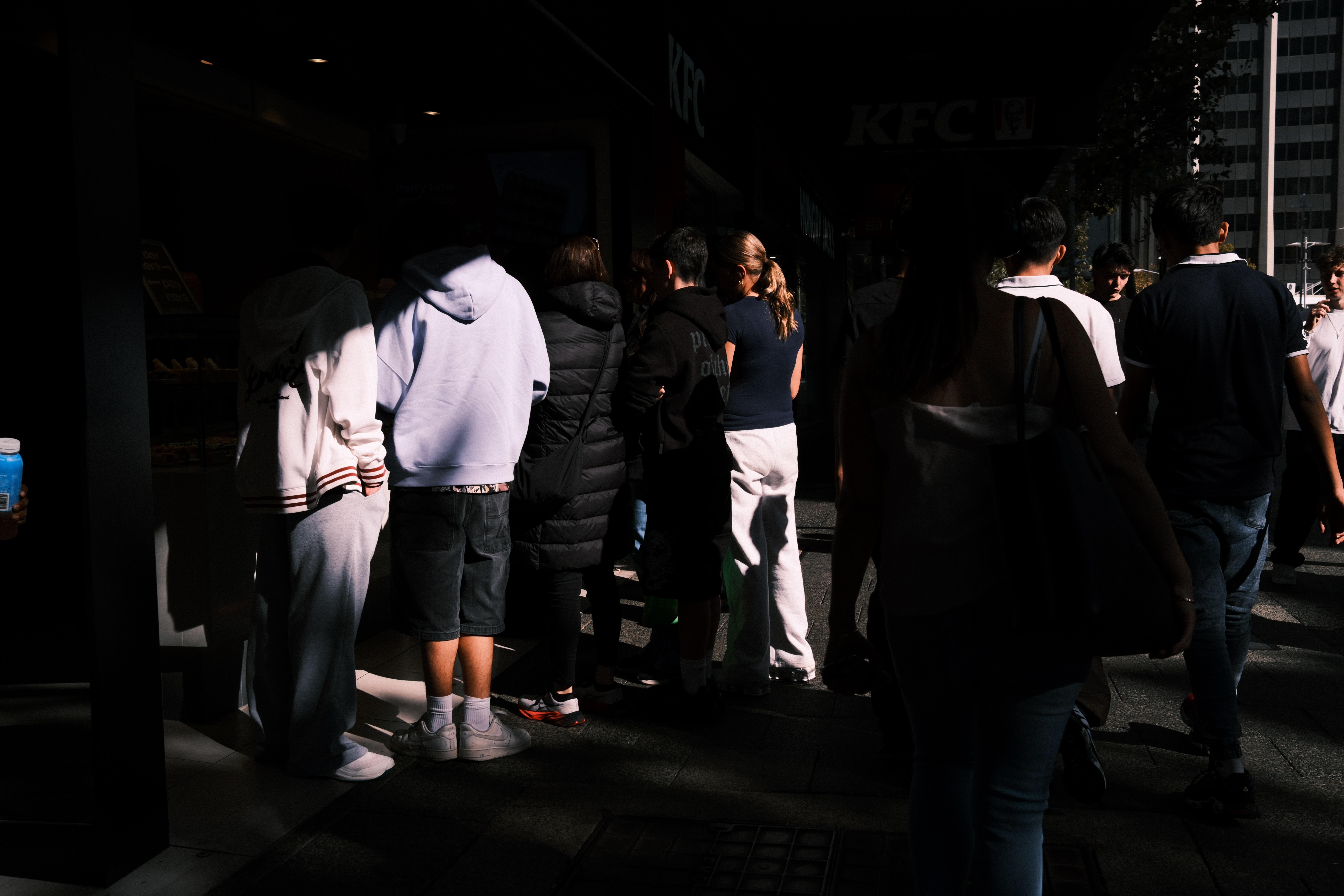 People standing in a line on a city sidewalk outside a storefront, with strong sunlight illuminating part of the group and deep shadows across the rest; pedestrians pass on the right and a tall building is visible in the background.