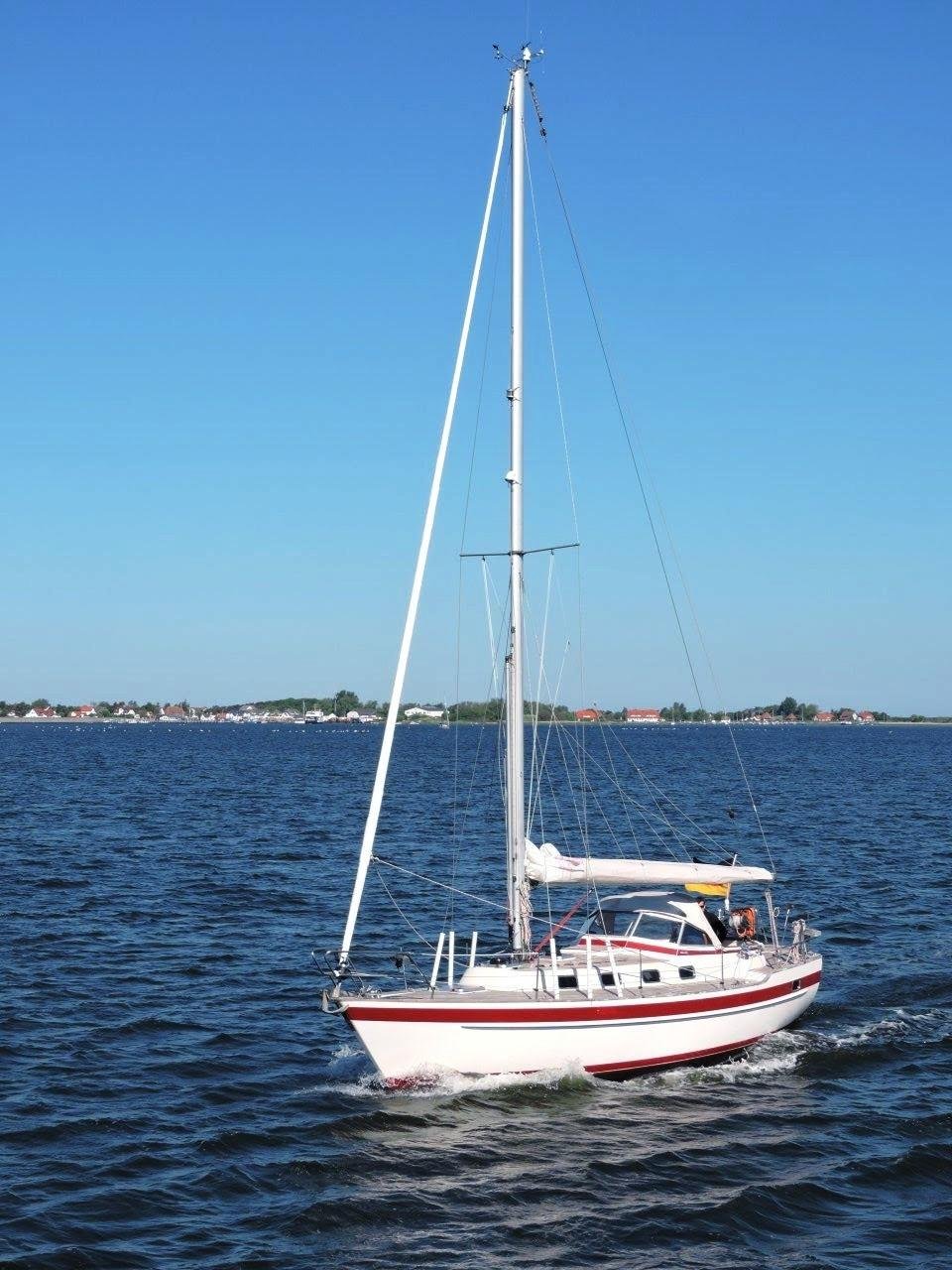 A white and red sailboat glides on a calm blue sea under a clear sky. The boat's sails are down, and a person is visible near the cockpit. In the background, a distant shoreline with buildings and greenery stretches across the horizon.