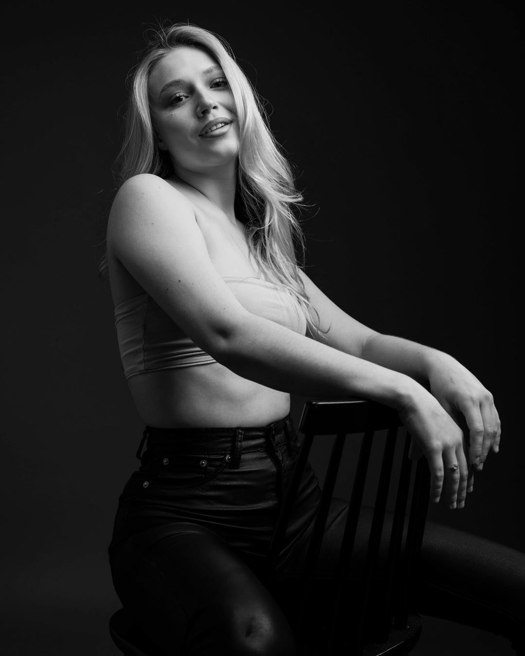This is a black and white photo of a person with long hair, sitting on a chair. The person is wearing a sleeveless top and high-waisted pants. They are looking slightly upwards and smiling.