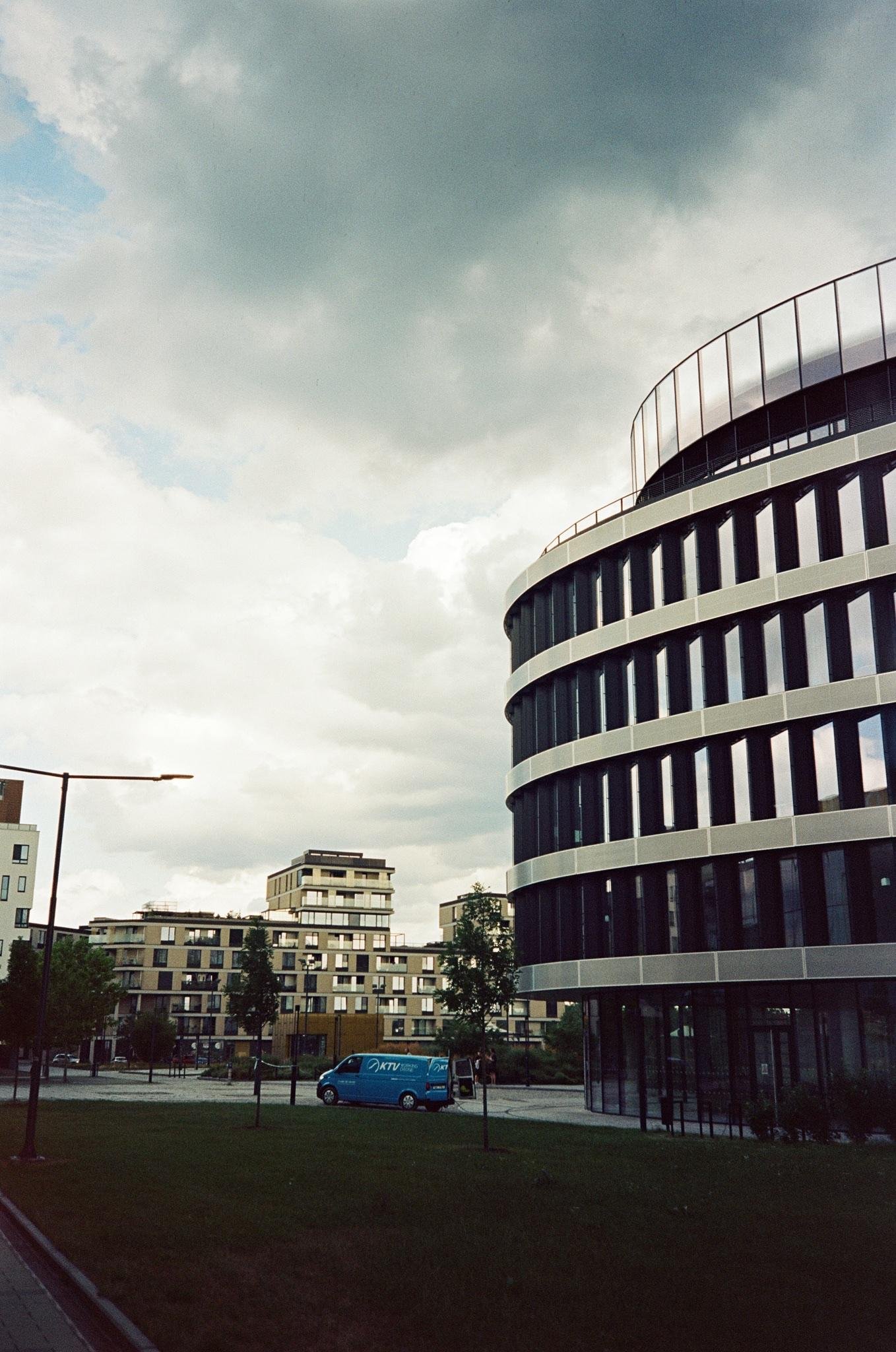 Curved modern office building on the right with vertical dark windows and horizontal concrete bands, viewed from street level; residential apartment blocks and trees in the background, a blue van parked near a walkway, and an overcast sky above.