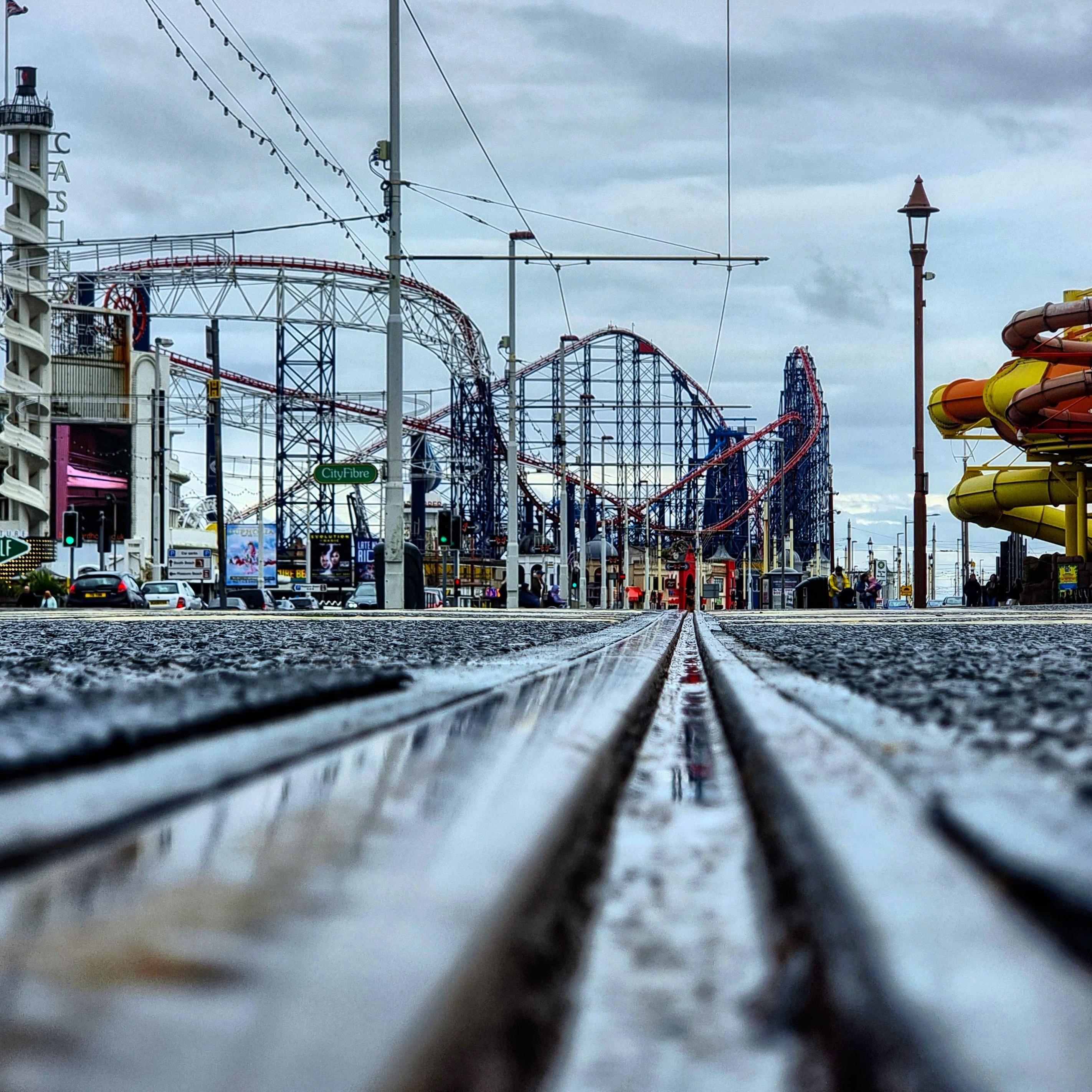 The image shows a low-angle view of tram tracks on a street with a roller coaster in the background. The scene includes buildings and streetlights, and there are some amusement park elements visible, like the roller coaster structure. The overall atmosphere suggests a city or town setting with recreational attractions.