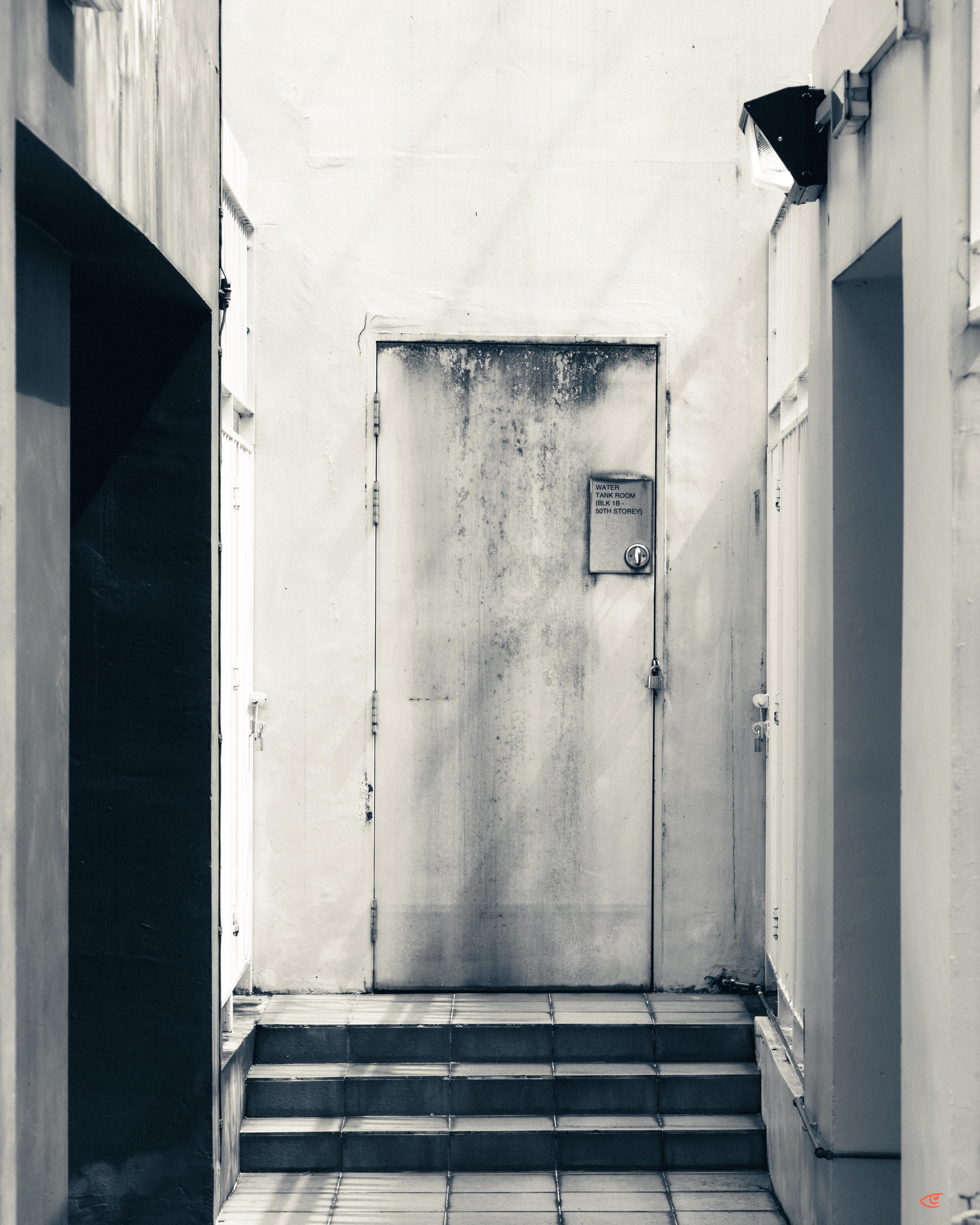 Stained metal door at the top of three tiled steps in a narrow corridor, with a small warning sign near the handle and plain walls and side openings on both sides.