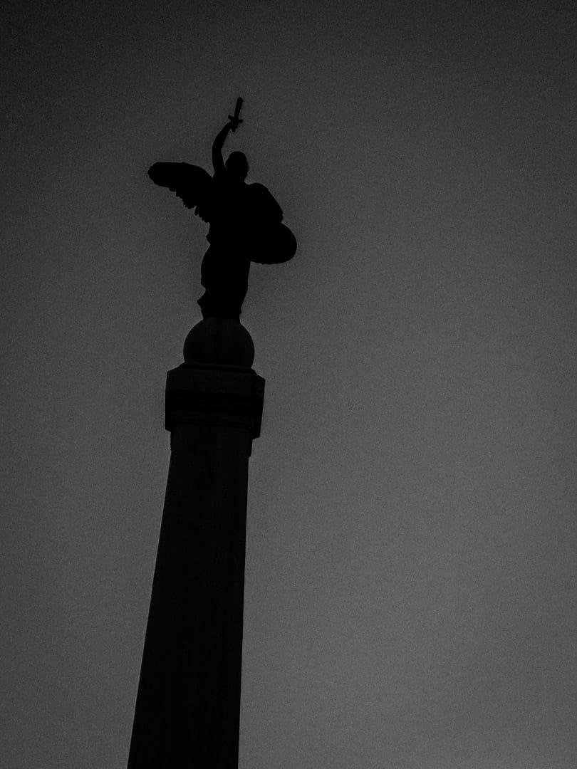 Silhouette di statua alata che regge un oggetto che potrebbe essere una torica o una spada spezzata, tenendo il braccio verso l'alto. Il soggetto è in silhouette e la foto è in bianco e nero.