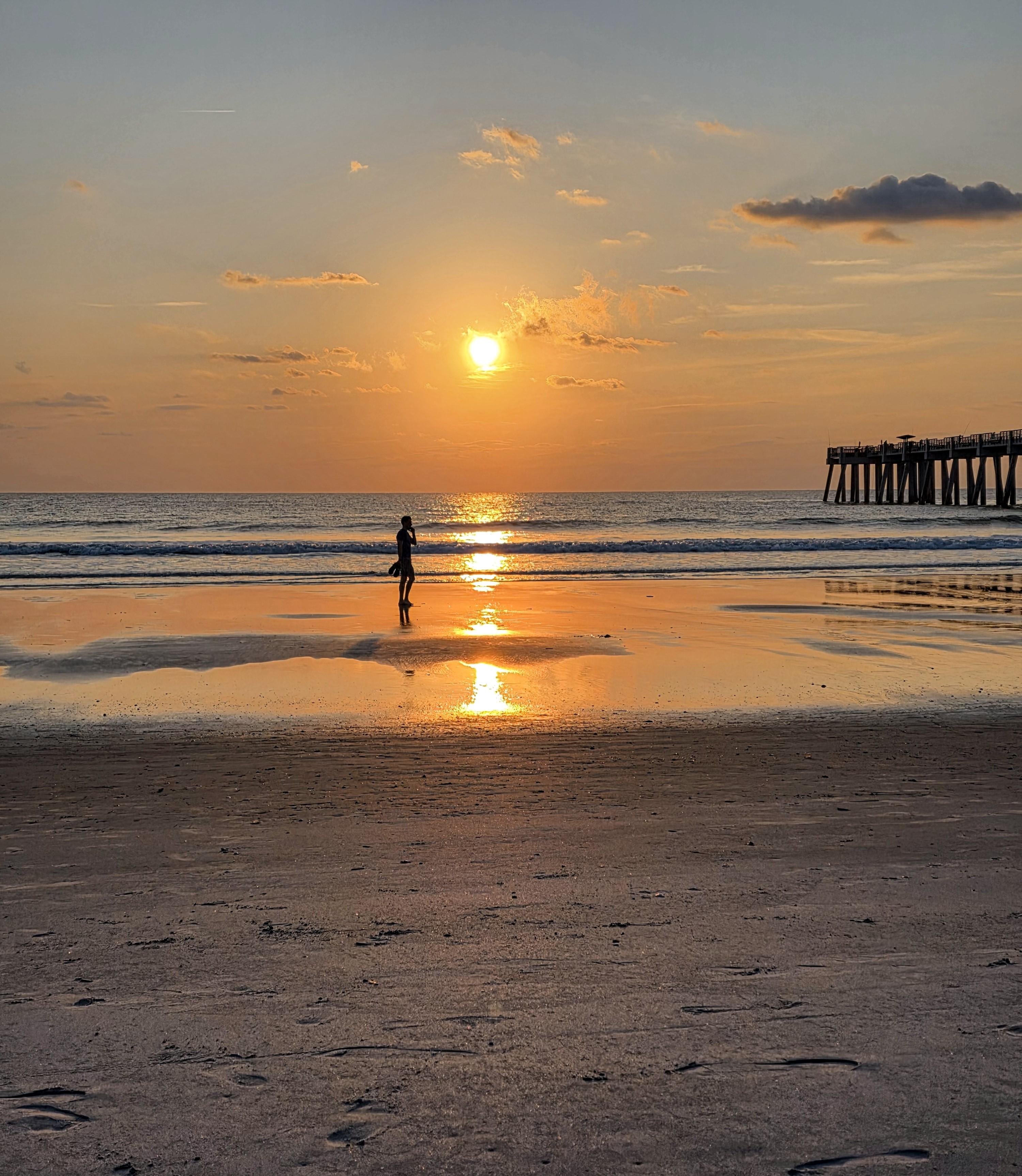 Sunris over a sandy beach with gentle waves. A person stands near the water's edge, silhouetted by the rising sun. The sky is filled with clouds, some of which are lit by the orange and gold hues of the sunrise. A pier extends from the right side into the ocean. Reflections of the sun on the wet sand create a glowing path.