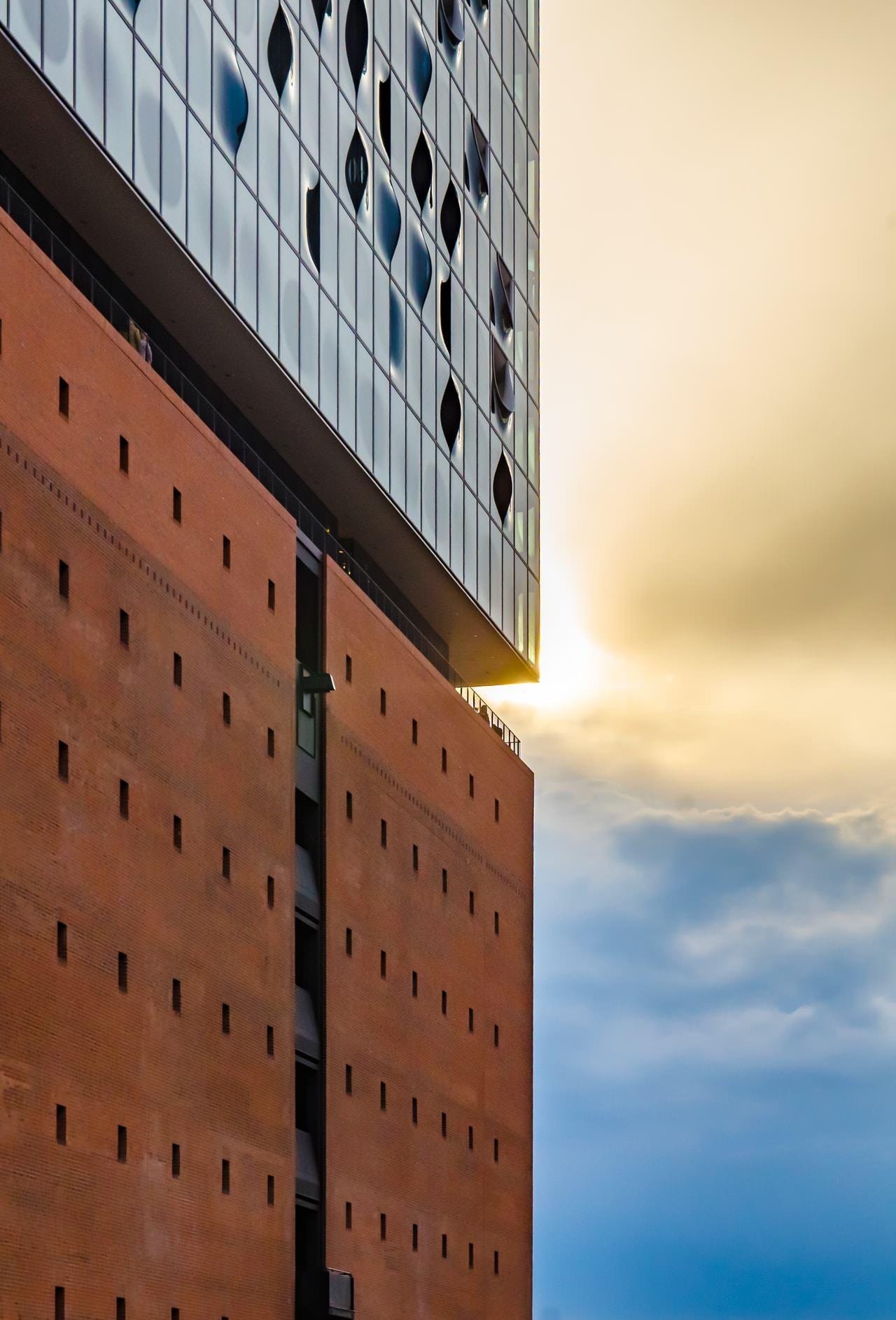 Low-angle view of a modern building with a tall brick base punctuated by small square openings and an overhanging glass upper section with diamond-shaped window cutouts, set against a partly cloudy sunset sky.