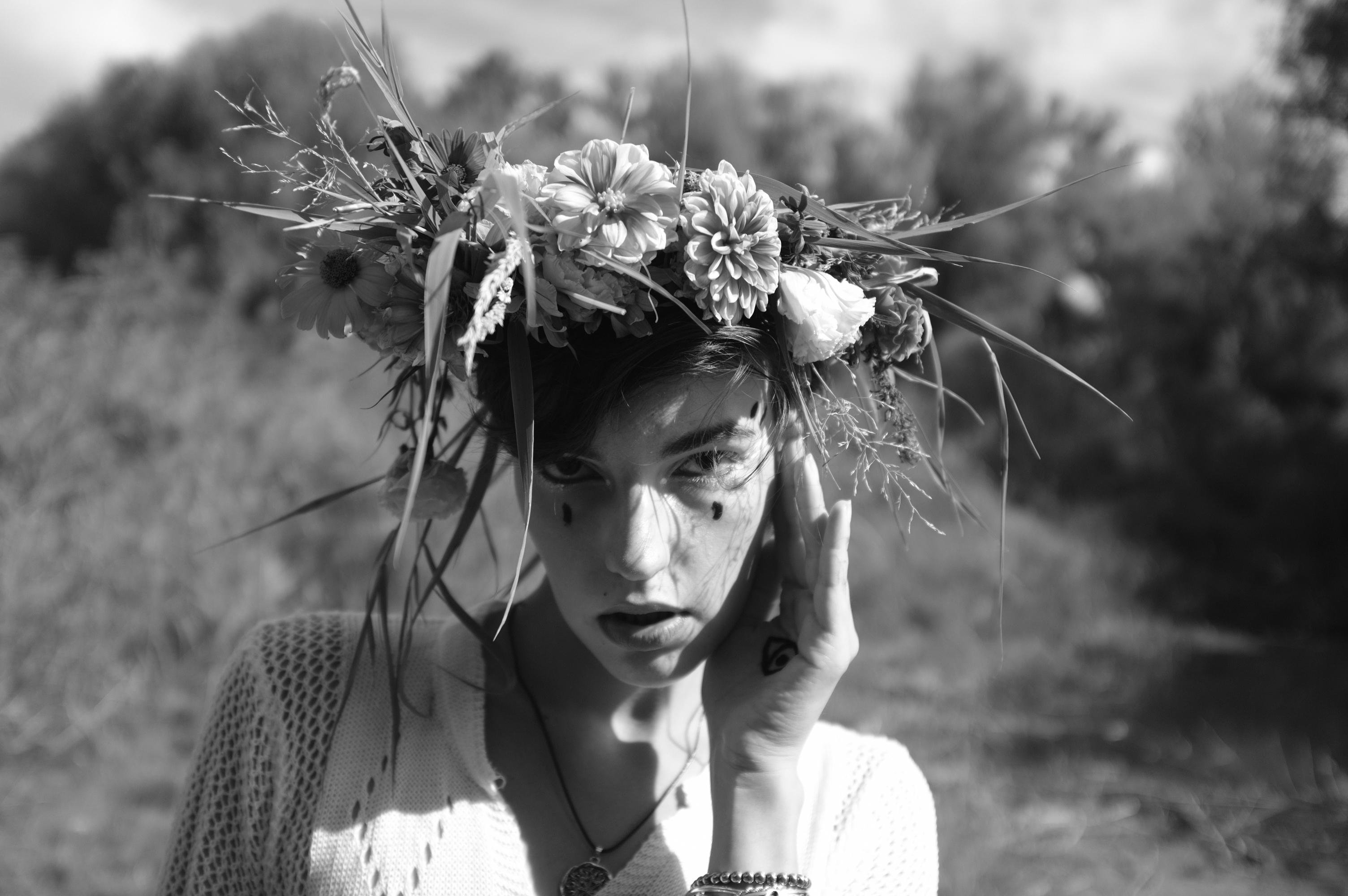 Black and white photo of a person wearing a floral crown made of daisies and other flowers, set against a blurred natural background. The person has short hair and wears a knitted top with a pendant necklace. They have hand-drawn teardrop marks under their eyes and rest one hand on their cheek, with a tattoo visible on the hand.
