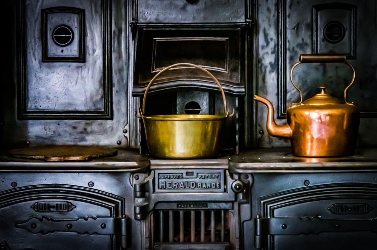 The image shows an antique cast iron stove, with a kettle and a pot on top. The stove has ornate details and the word "Herald Range" is visible. The kettle and pot appear to be made of copper or brass, adding a vintage touch to the scene.